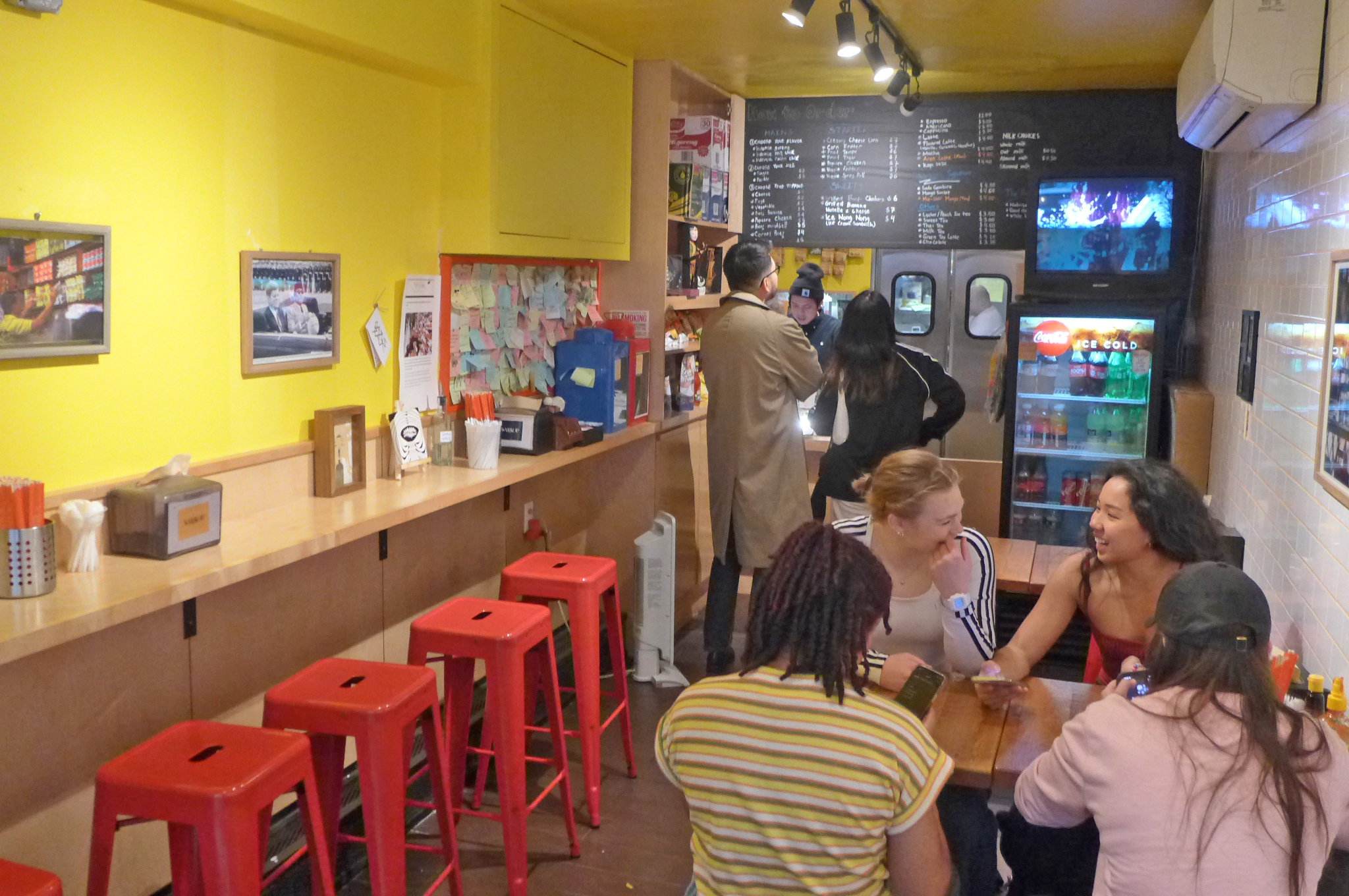 A yellow room with a table of laughing diners and an order counter at the rear with chalkboard menu.