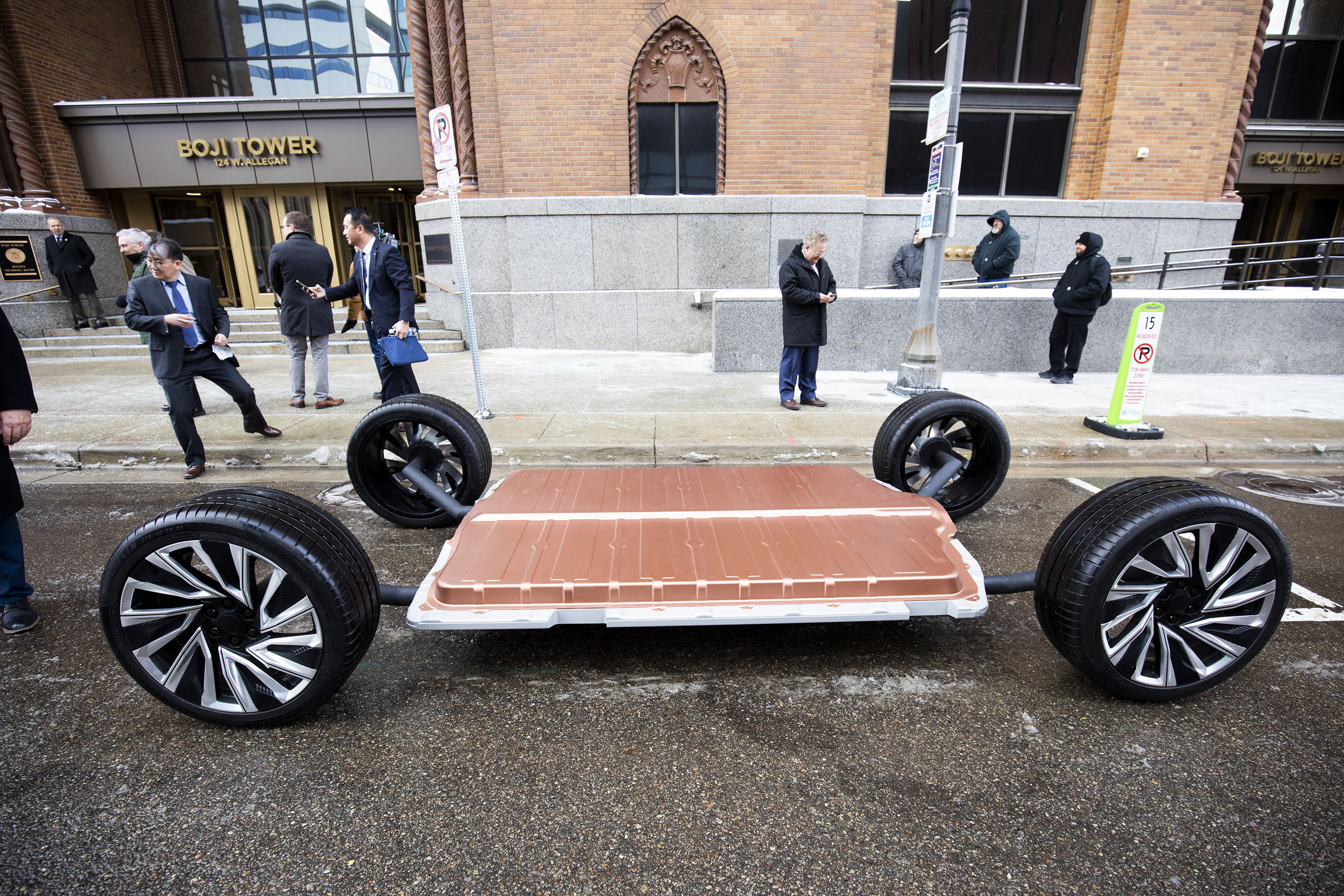 The chassis of an electric vehicle, which consists of a large rectangular battery connected to four wheels, sits on a road in front of a building.