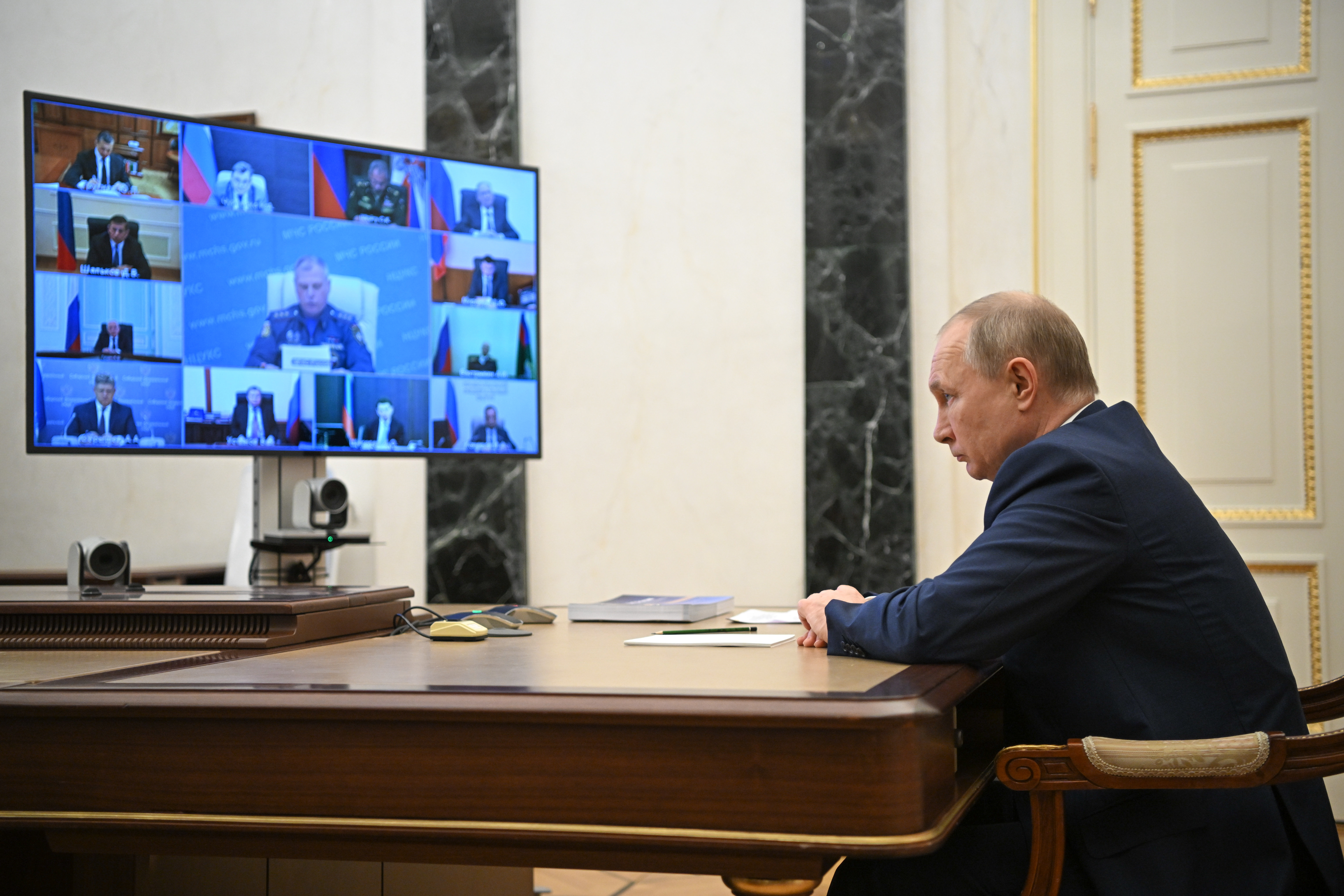 Putin sitting across from a large screen that shows several participants in the meeting.