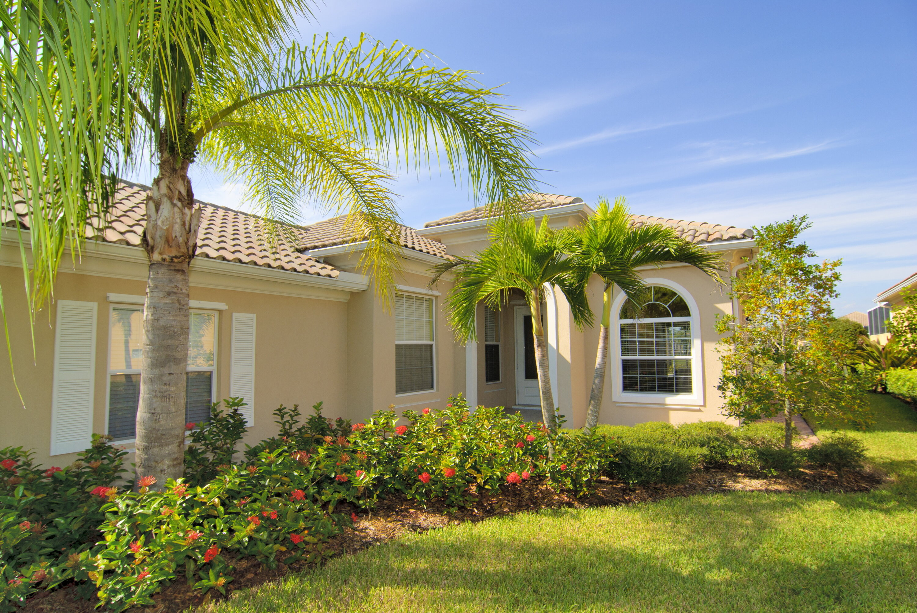 A beige one-story Floridian house with a light brown tiled roof, a well manicured lawn with tropical flower bushes and small palm trees