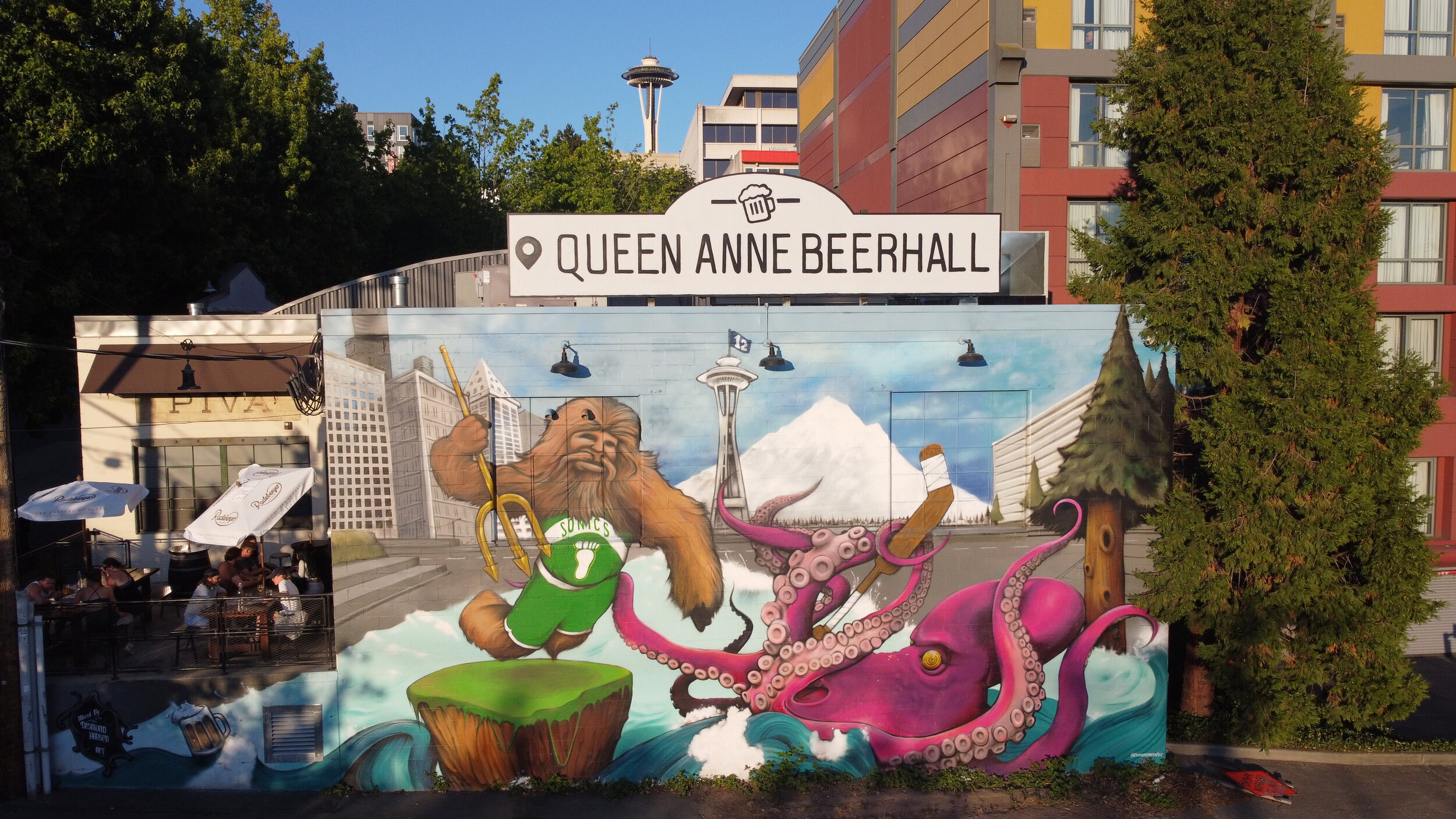 A mural on the exterior of Queen Anne Beerhall depicts Sasquatch attacking a kraken. Above it is the restaurant’s sign. To the left side, patio seating is visible.