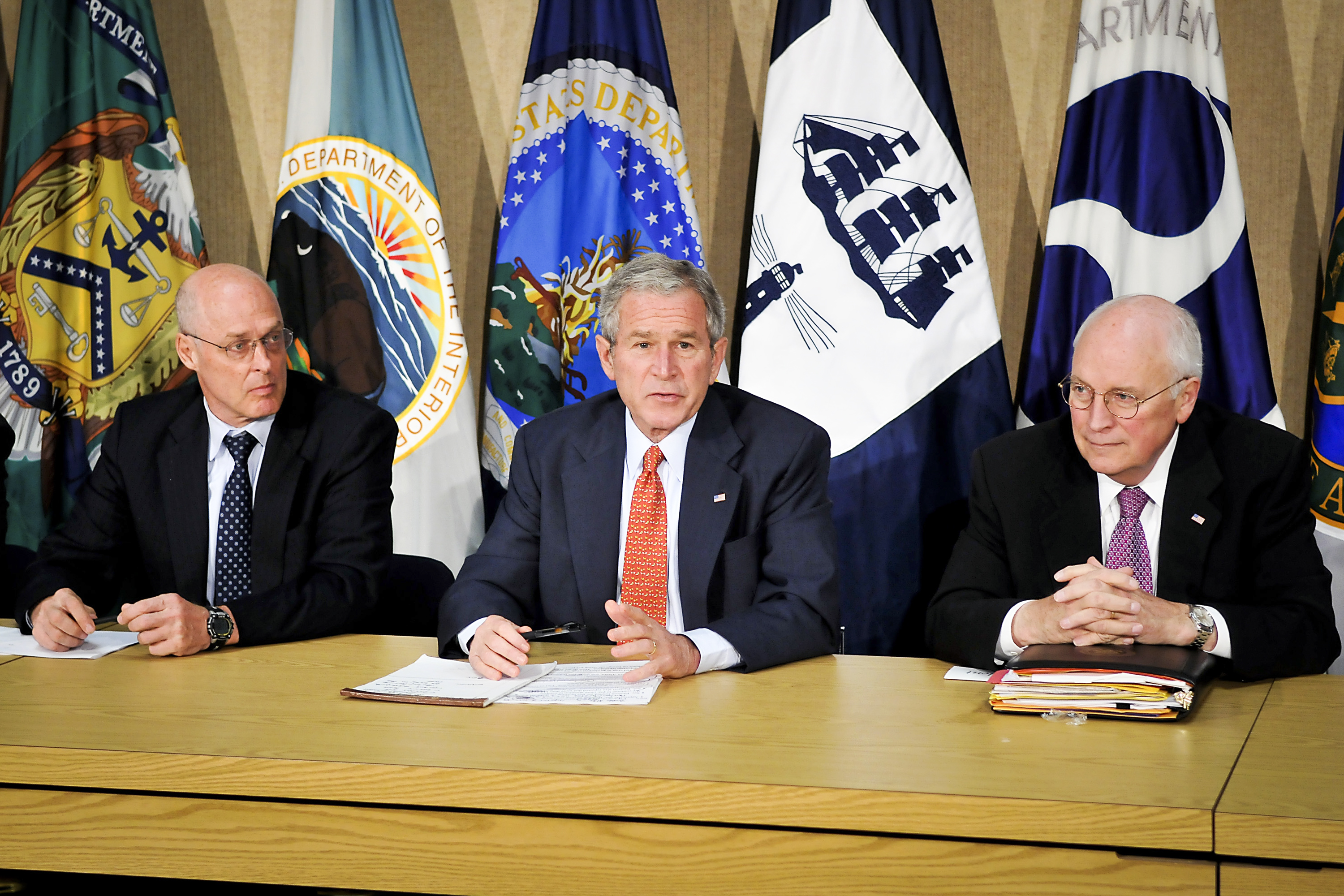 Three politicians sitting in a row at a table in front of multiple flags.