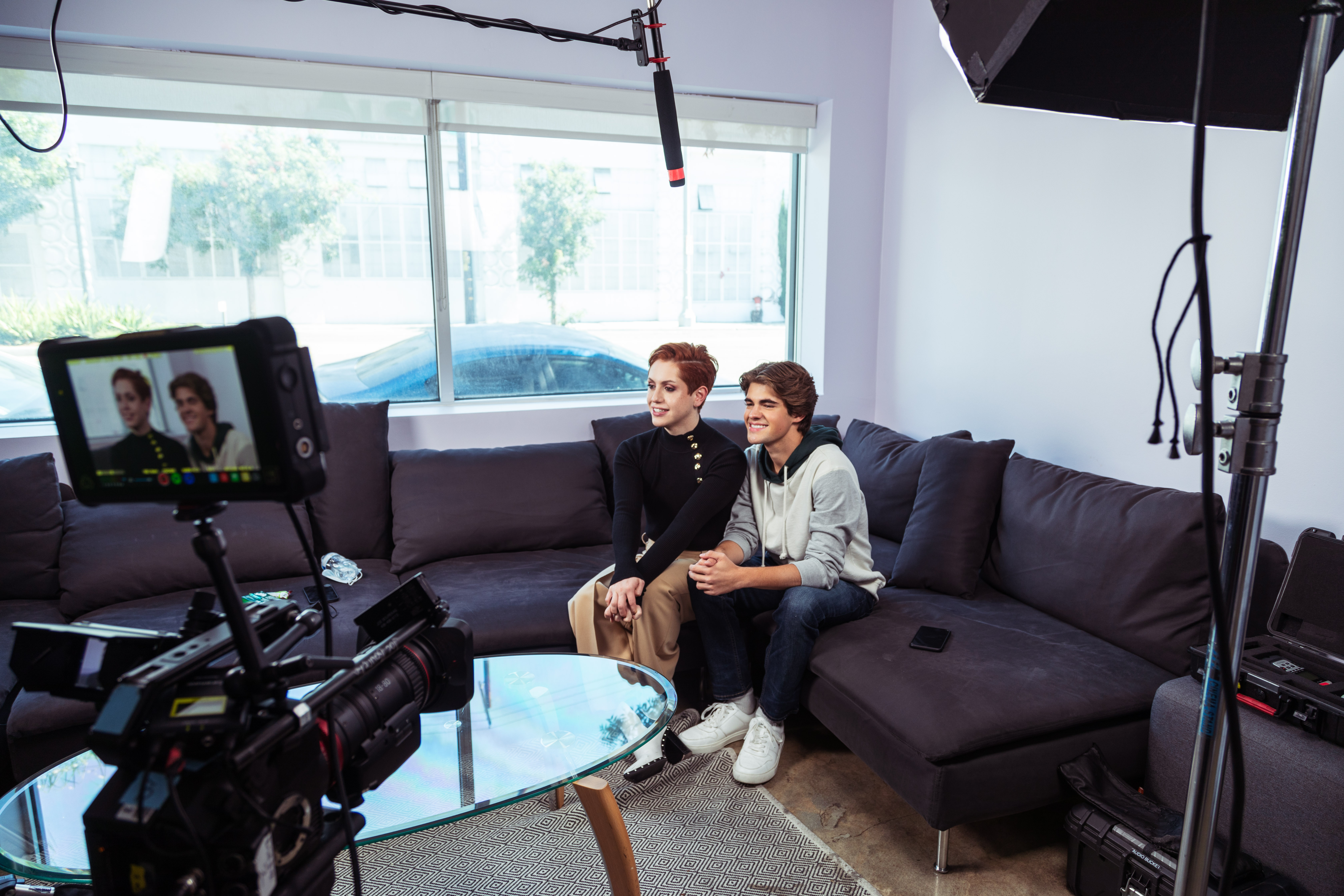 Two actors sitting on a studio couch being filmed.