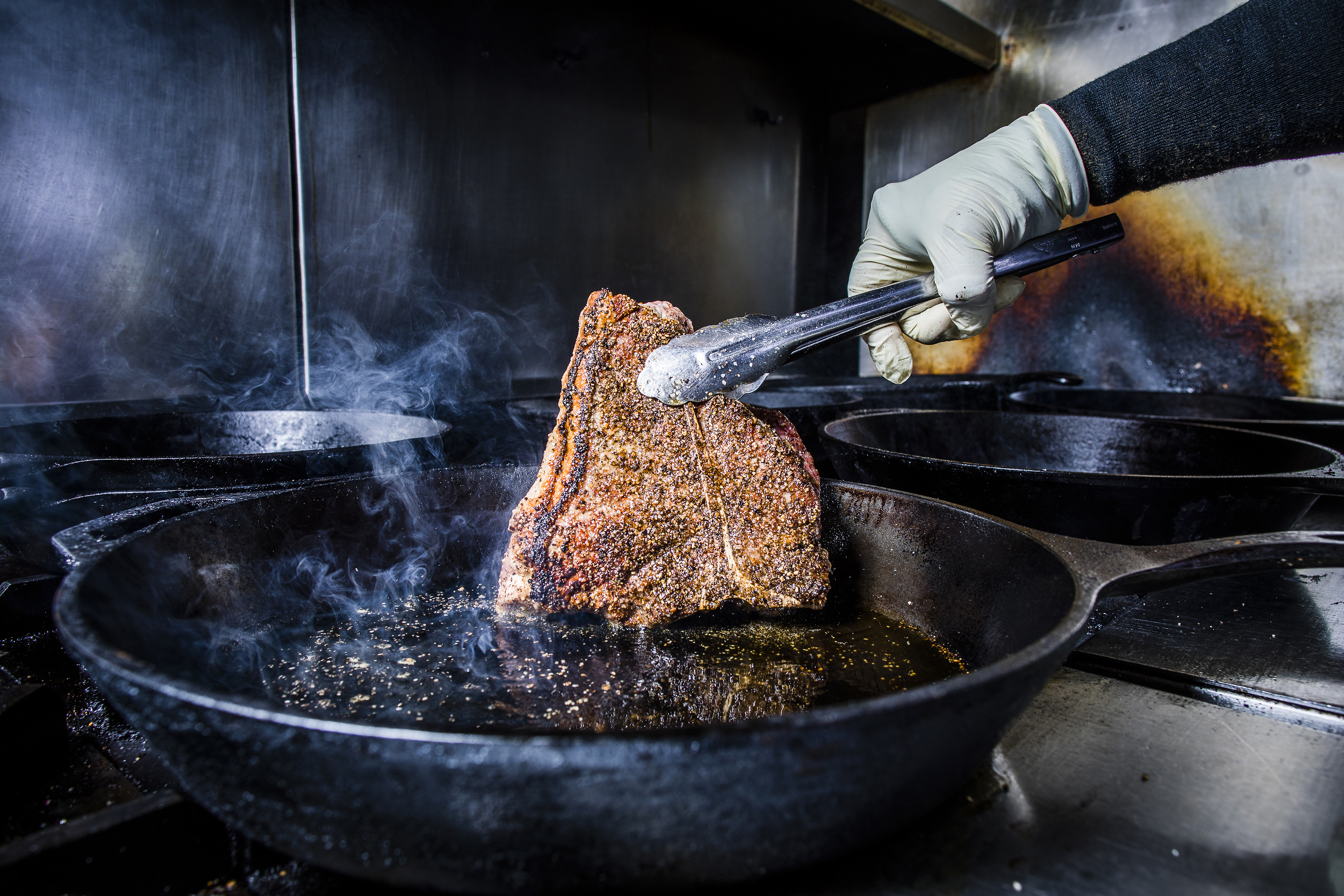 A hand gripping tongs that are holding a steak that is searing over a heated cast iron pan.