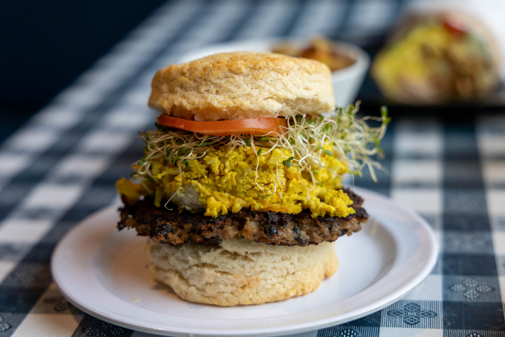 A biscuit sandwich on a white plate and a checked  table cloth stuffed with sprouts, tomator, and a plant-based sausage patty.