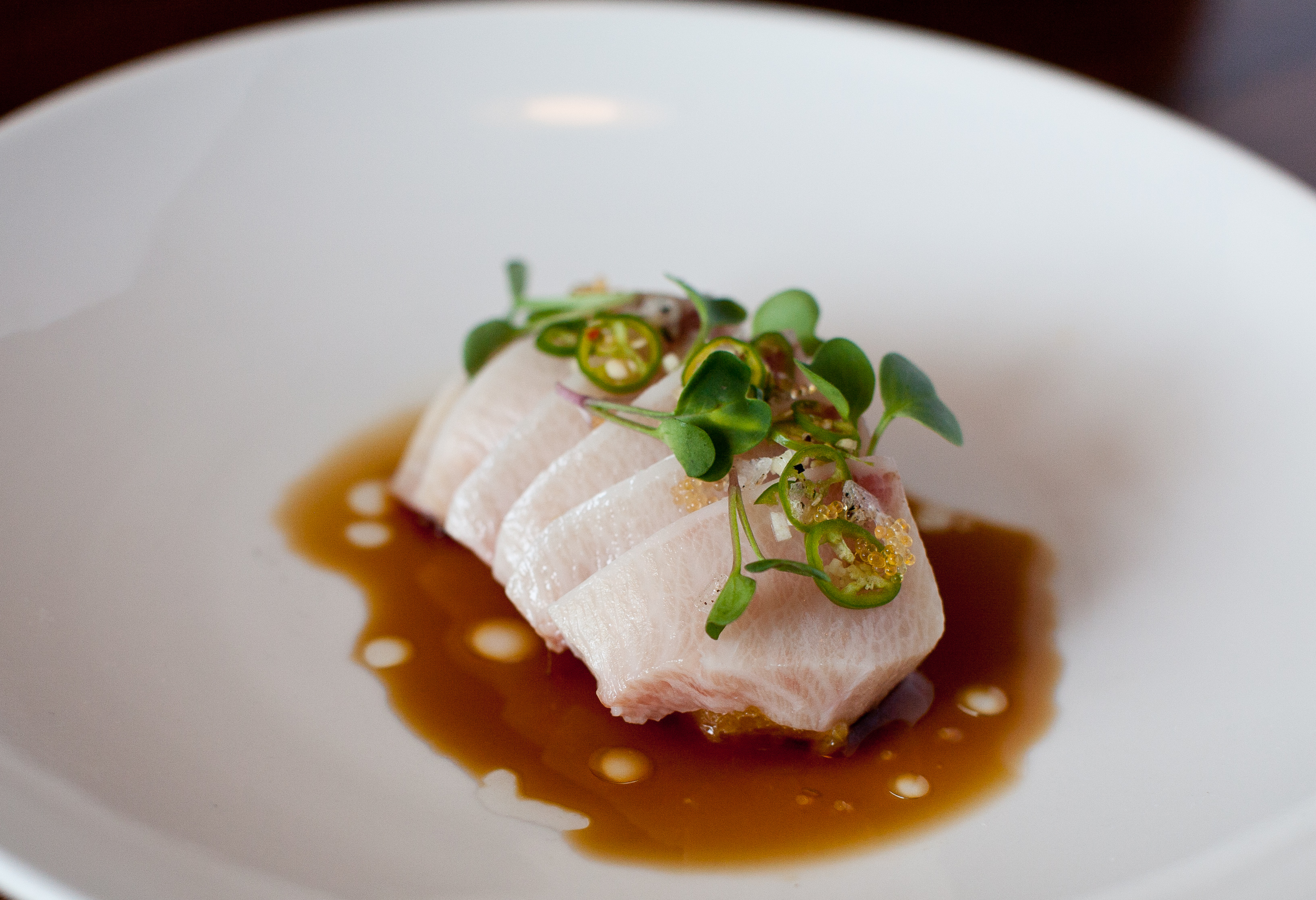 A dish with a piece of sliced-up light pink fish on a brown sauce topped with greens. 