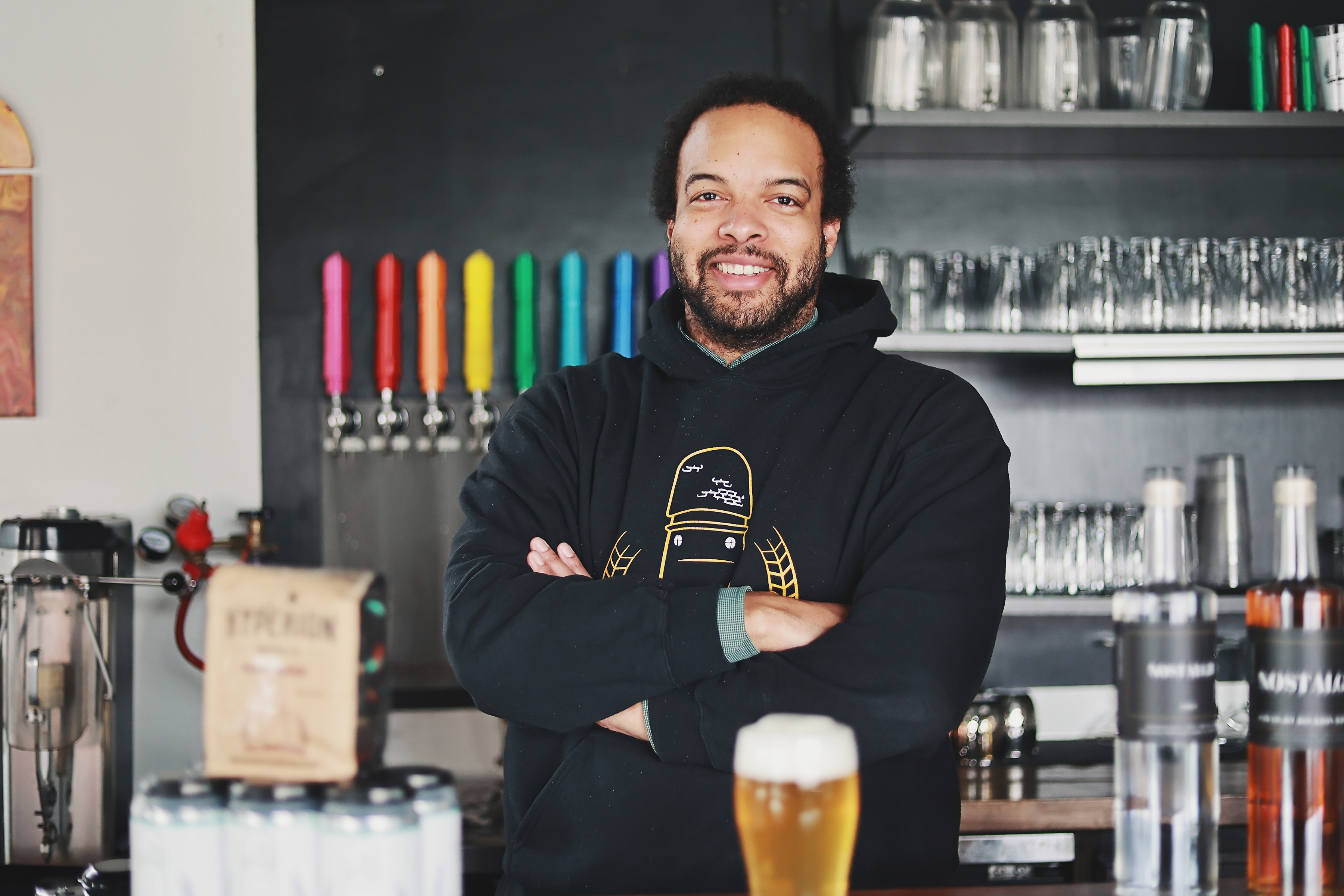 Brian Jones-Chance wears a black hooded sweatshirt with a cartoon of the Ypsilanti water tower on it and smiles behind the bar at 734 Brewing with colorful taps and a foamy, amber beer.