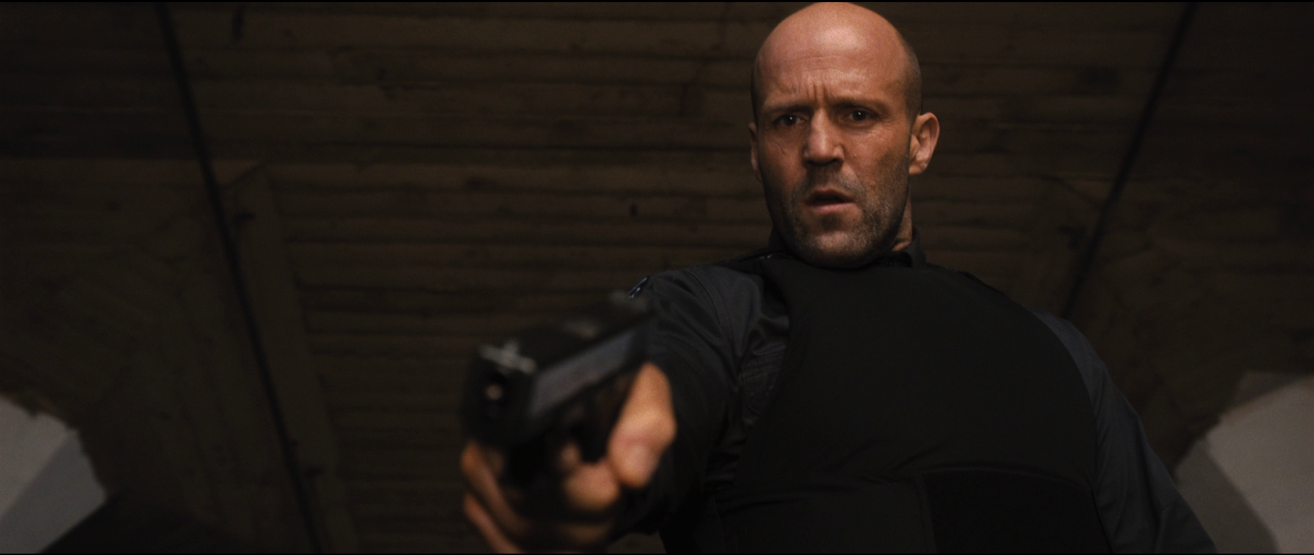 Jason Statham, wearing a bulletproof vest, points a pistol towards the camera in Wrath of Man.