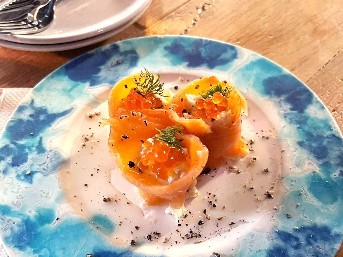 Three pieces of raw salmon are presented on a plate with a cloud-like blue rim, wrapped around roe and cream cheese.