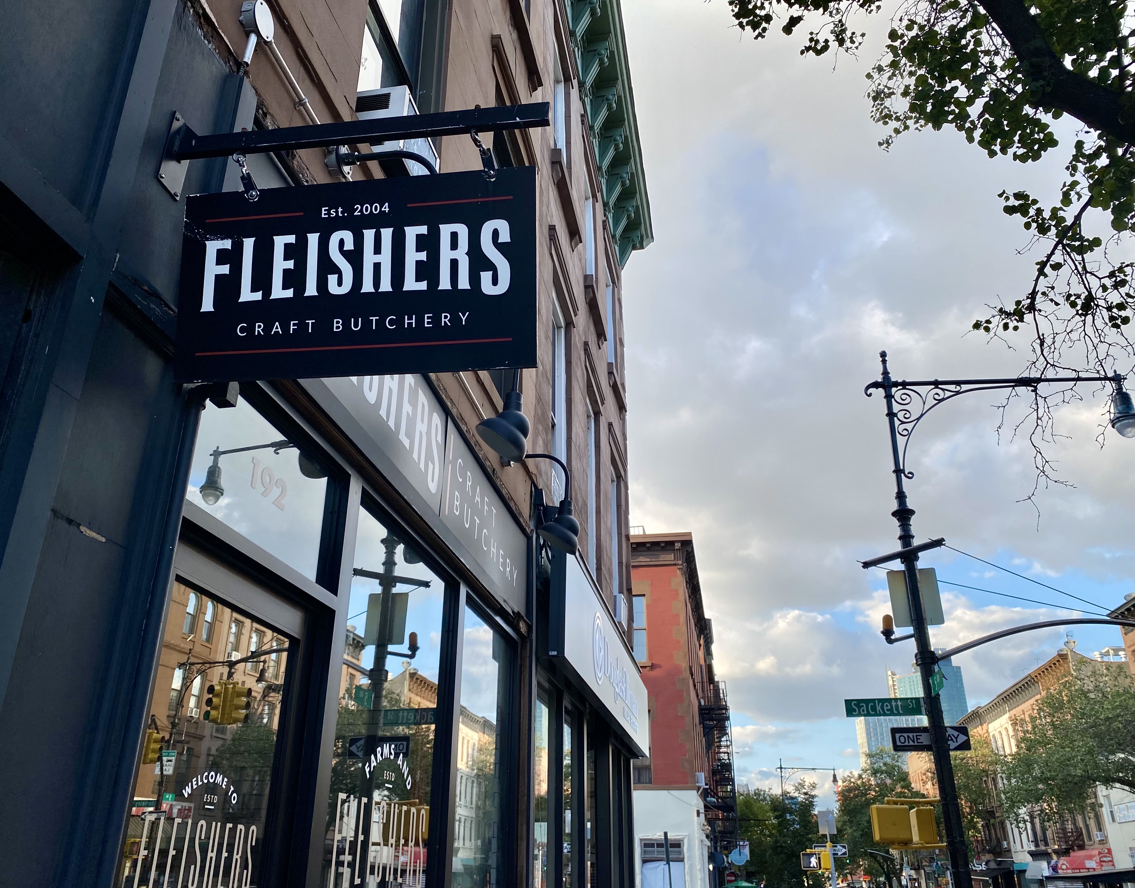 A black sign with the words “Fleishers Craft Butchery” hangs in a city street. In the background a storefront and clouds in the sky are visible.