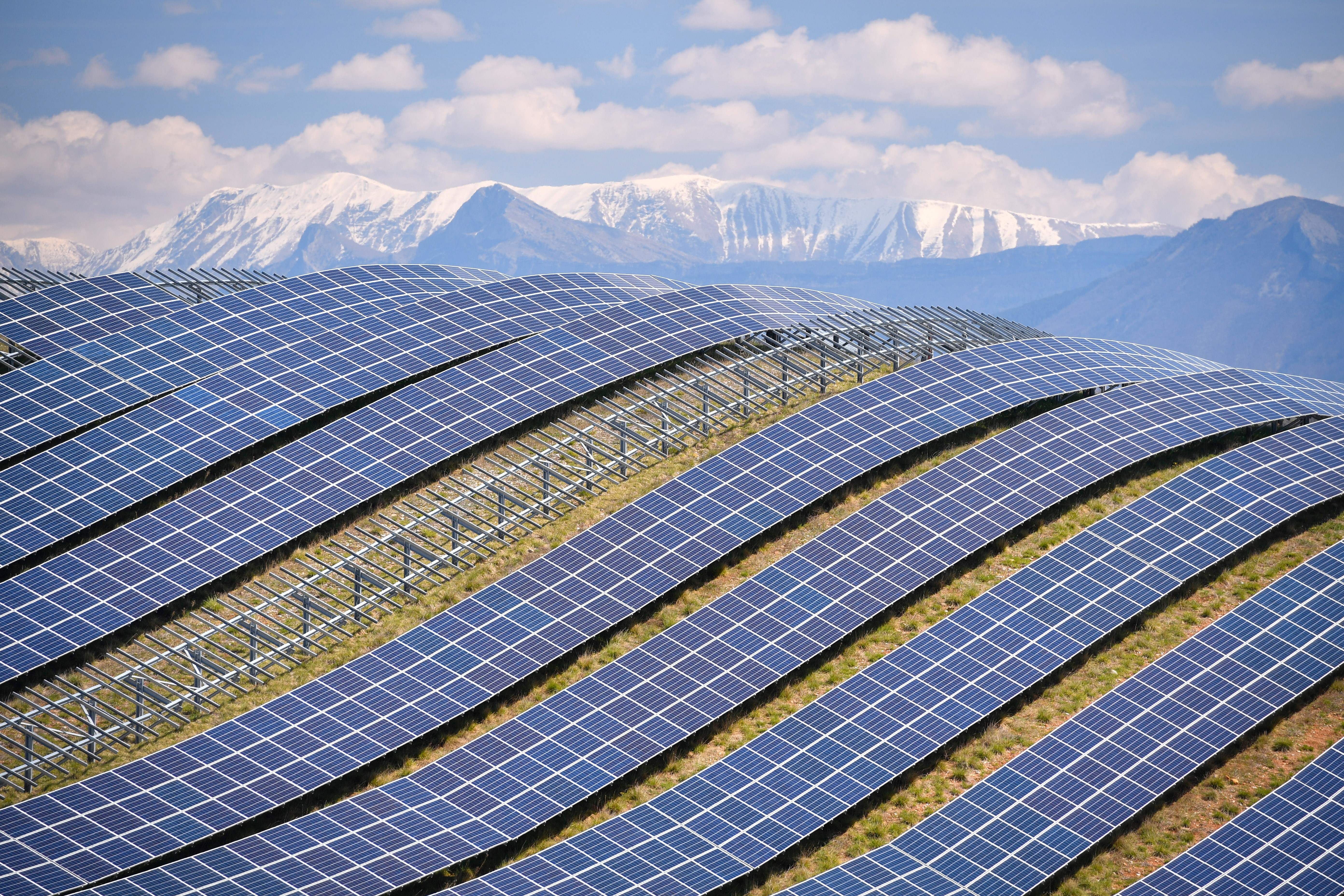 Long lines of solar panels follow the curve of a hillside, with snowcapped mountains in the distance.