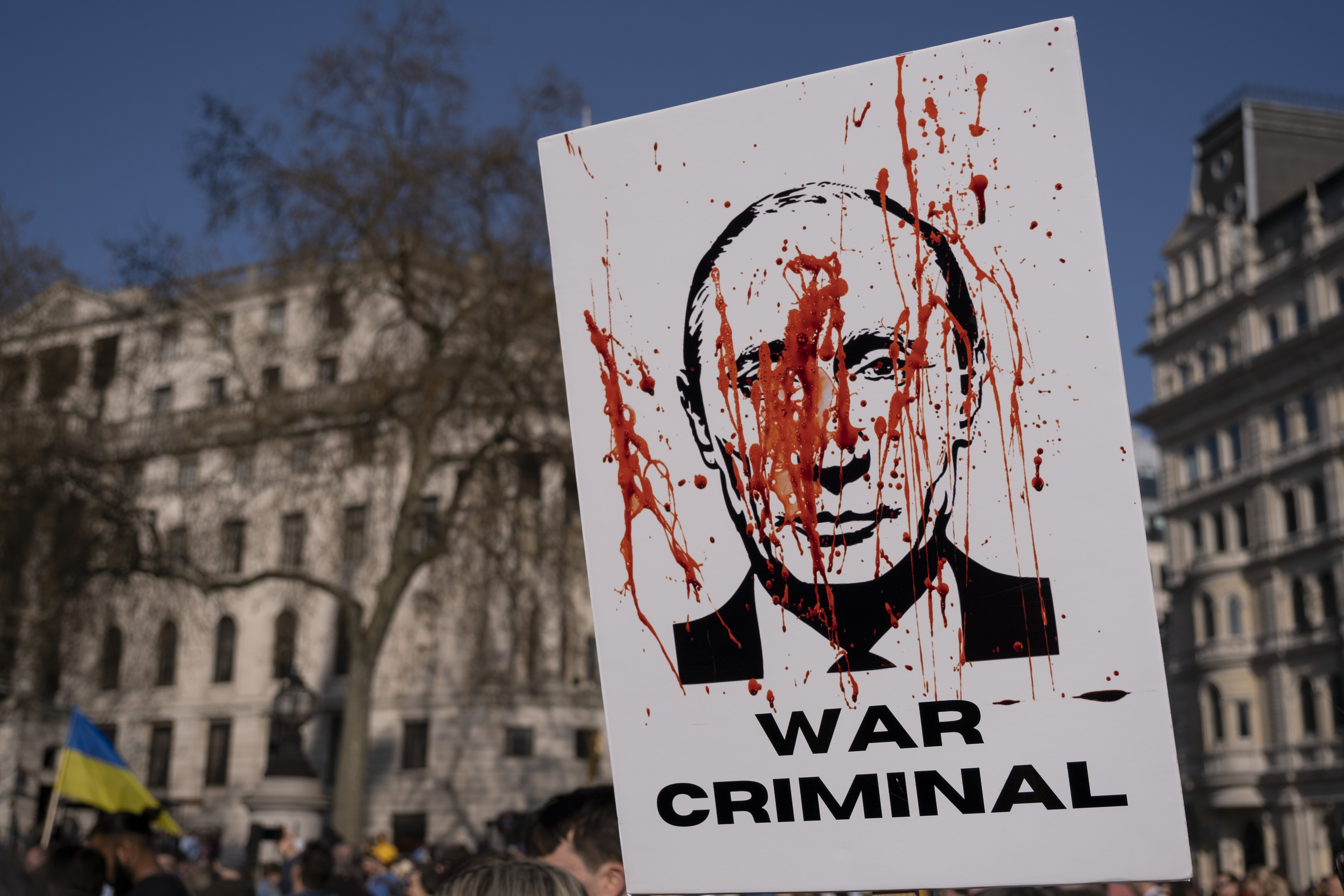 White sign with image of Russian President Vladimir Putin in black splattered in fake red blood with text that reads “war criminal.”