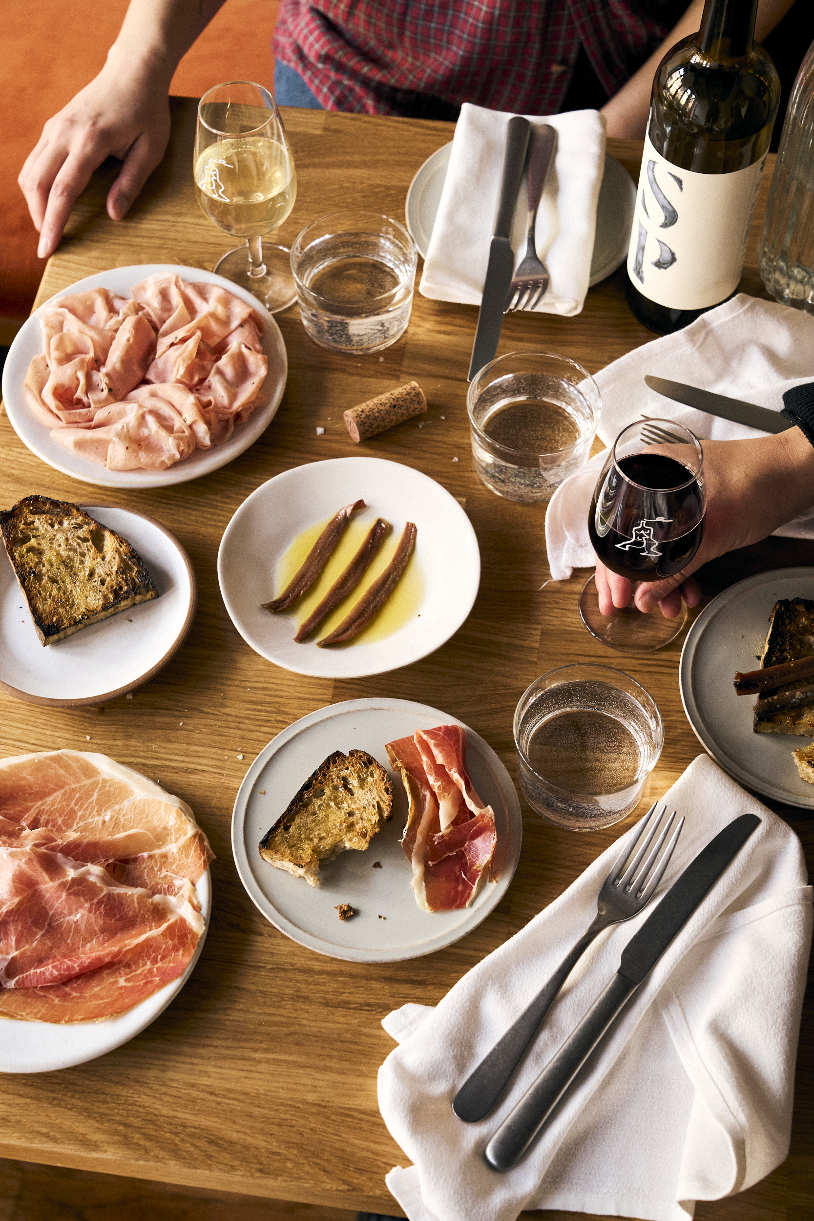 A wooden table with a spread of ham, mortadella, bread, anchovies, and glasses of wine.