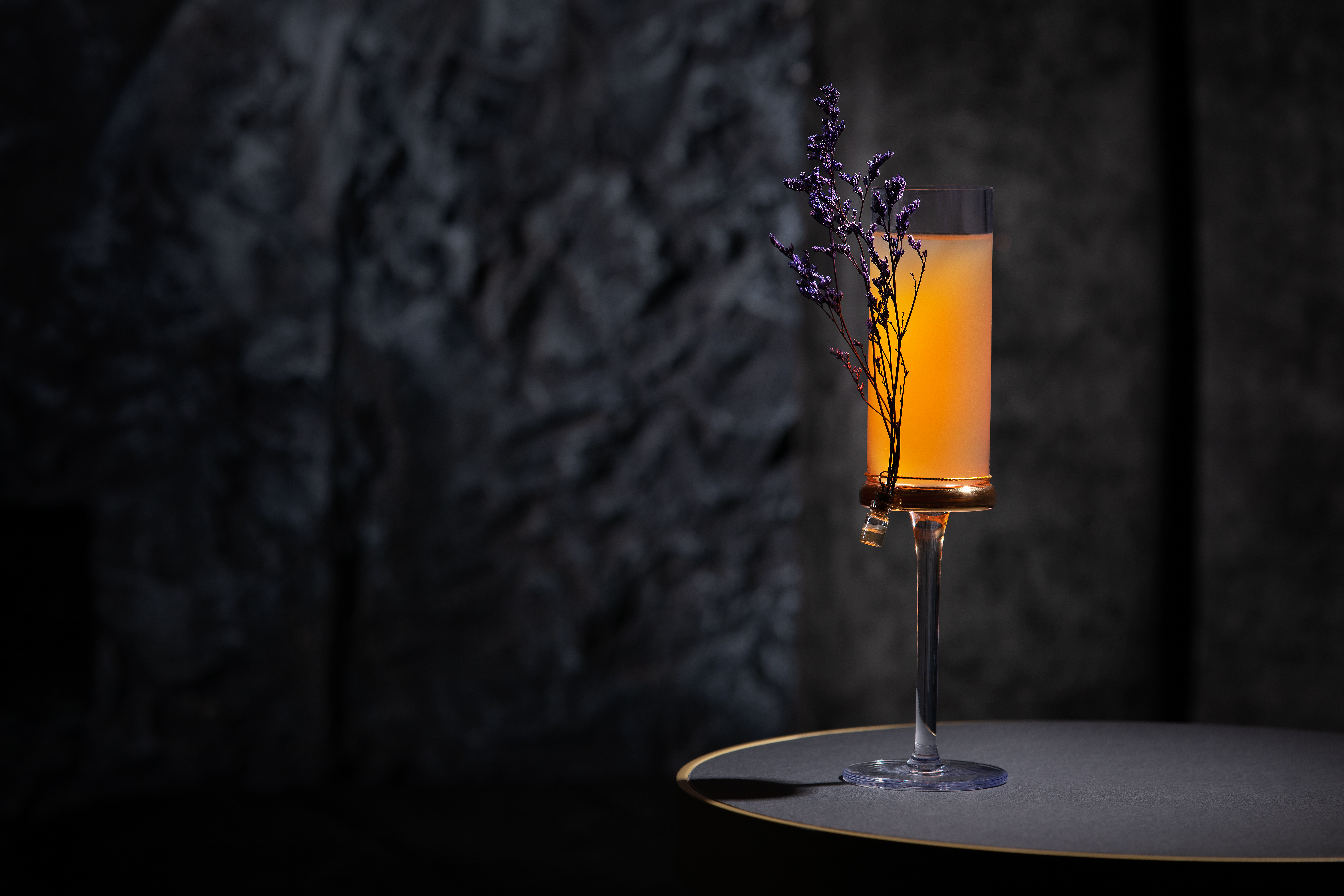 An orange cocktail is in a tall, narrow glass, embellished with a branch, photographed in front of a dark background.