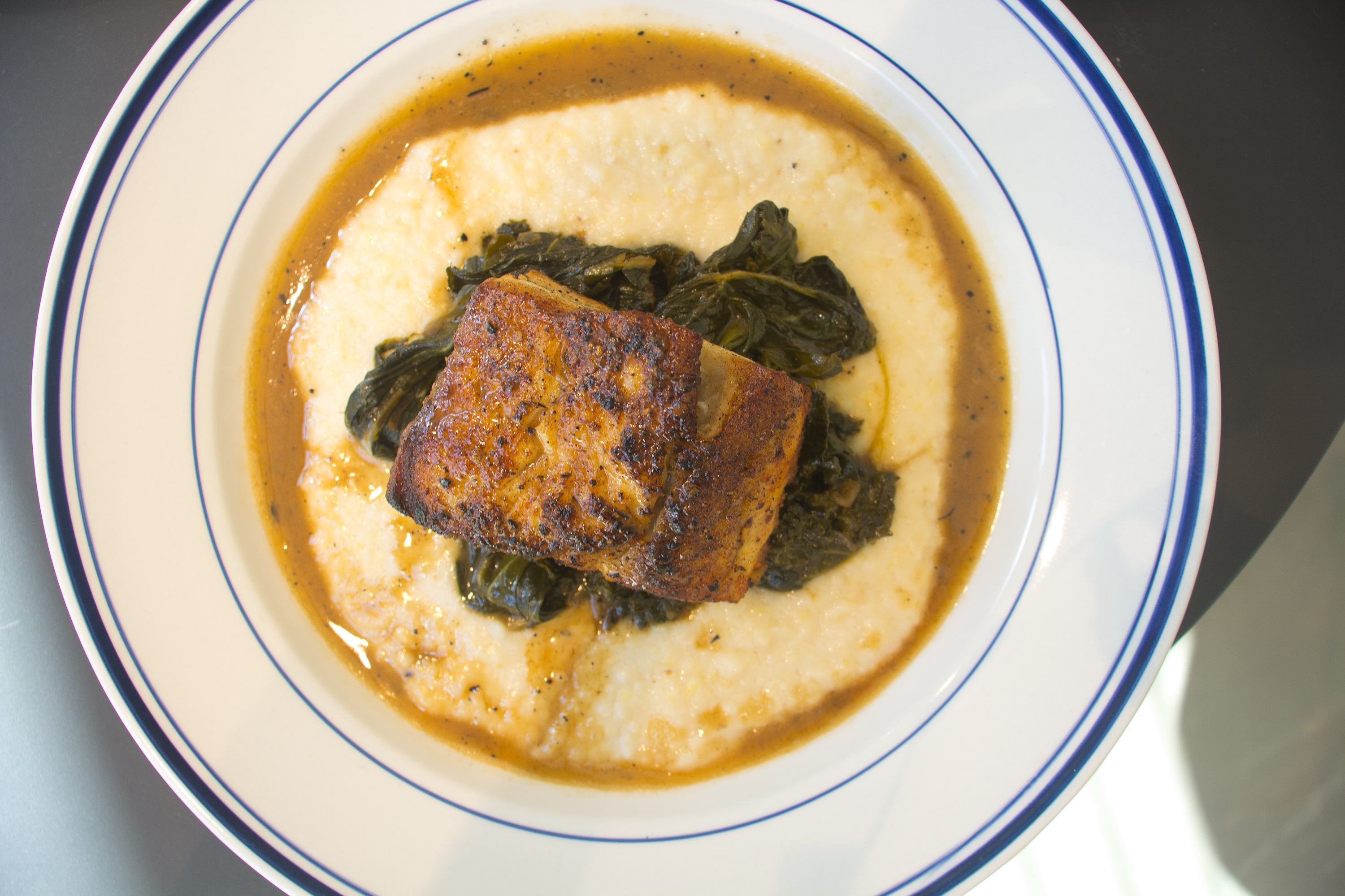 A plate of creamy grits in a brown sauce topped with cooked greens and a piece of brownish fish.