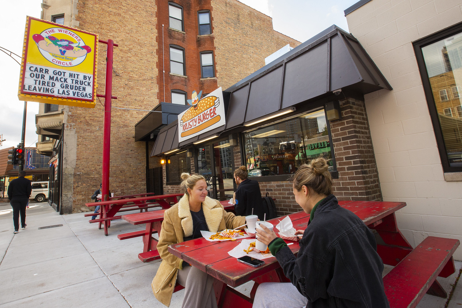 Two folks sitting on red picnic tables outside a hot dog stand with a tall sign in the background for Wiener’s Circle.