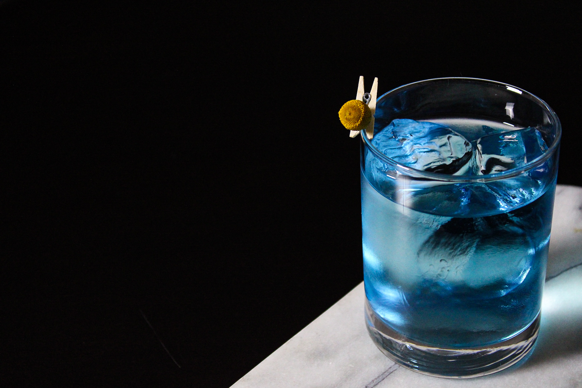 A clear blue cocktail sits on a white marble bar on a black background.