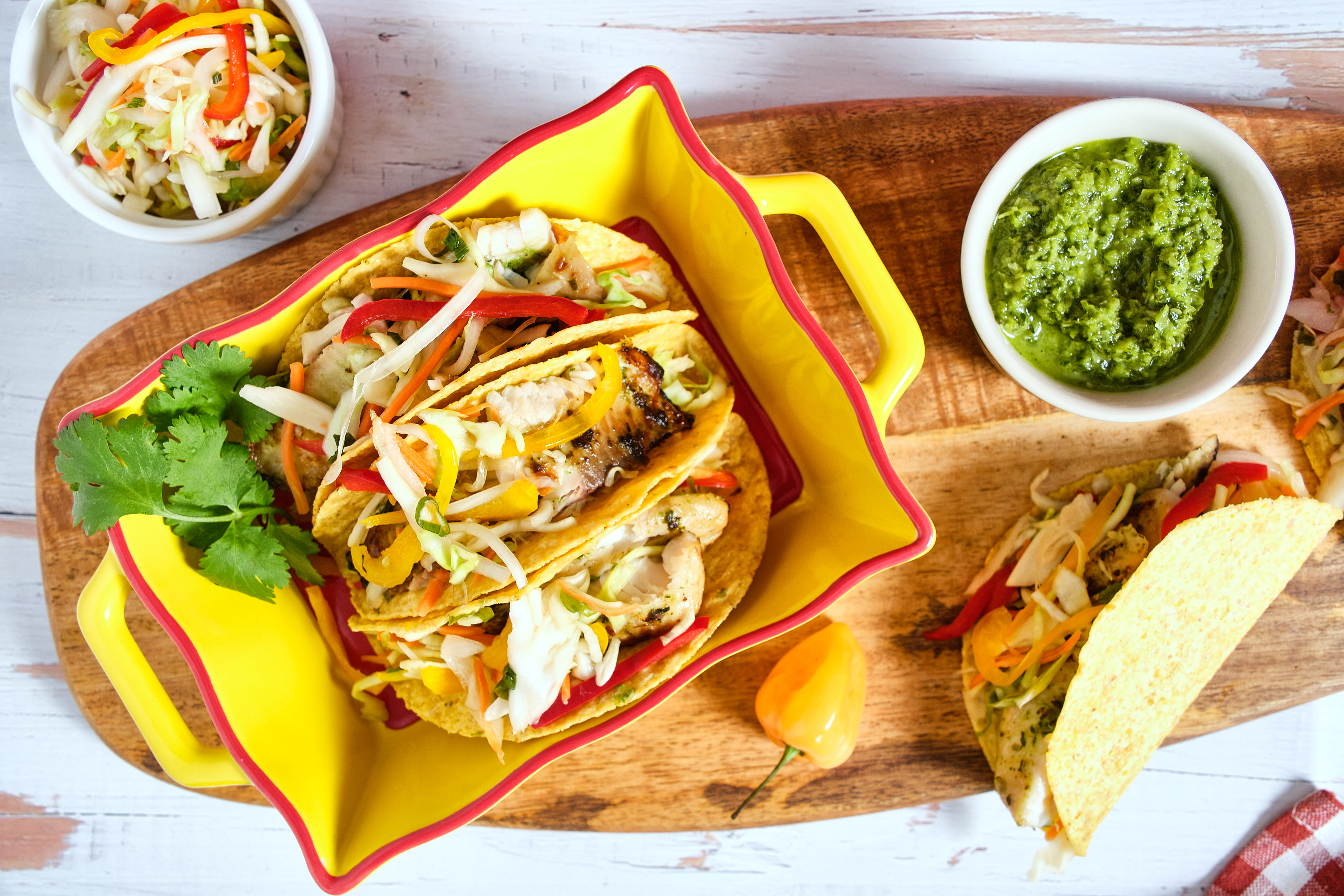 A plate of fish tacos on a wooden cutting board, served with bowls of pikliz and epi salsa