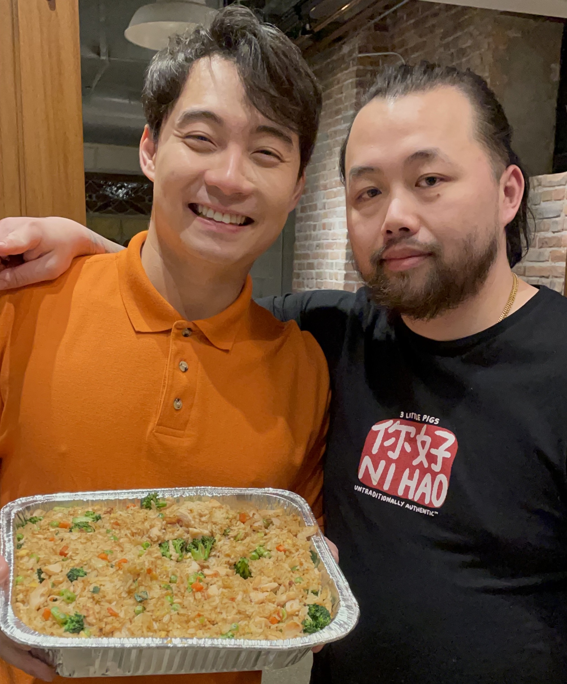 Two folks posing next to each other smiling, with one with an arm around another, with another holding a big tin of fried rice.