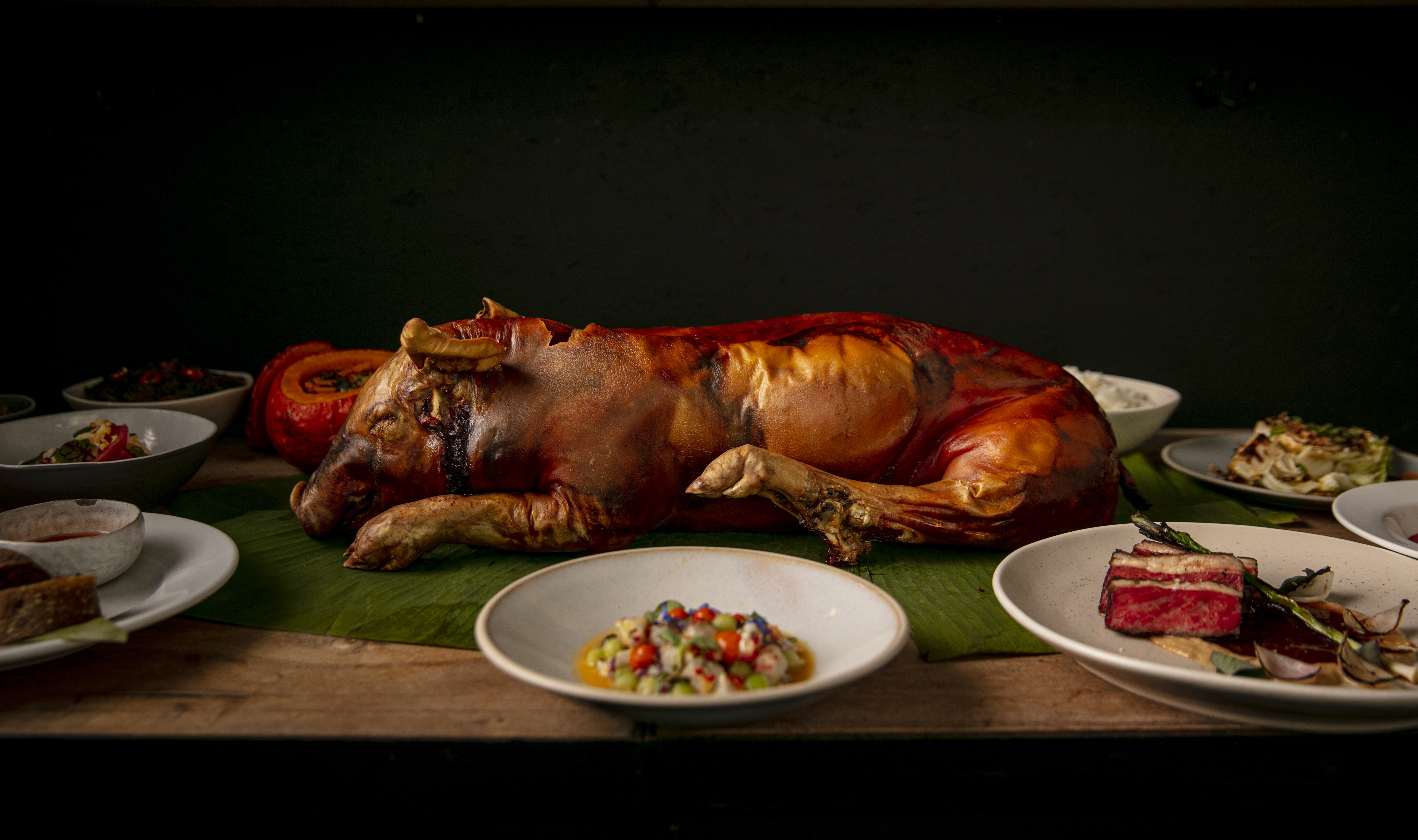 A suckling pig, stuffed with adobo rice, on a table surrounded by more Filipino dishes, front: kinilaw; to the right: bistek
