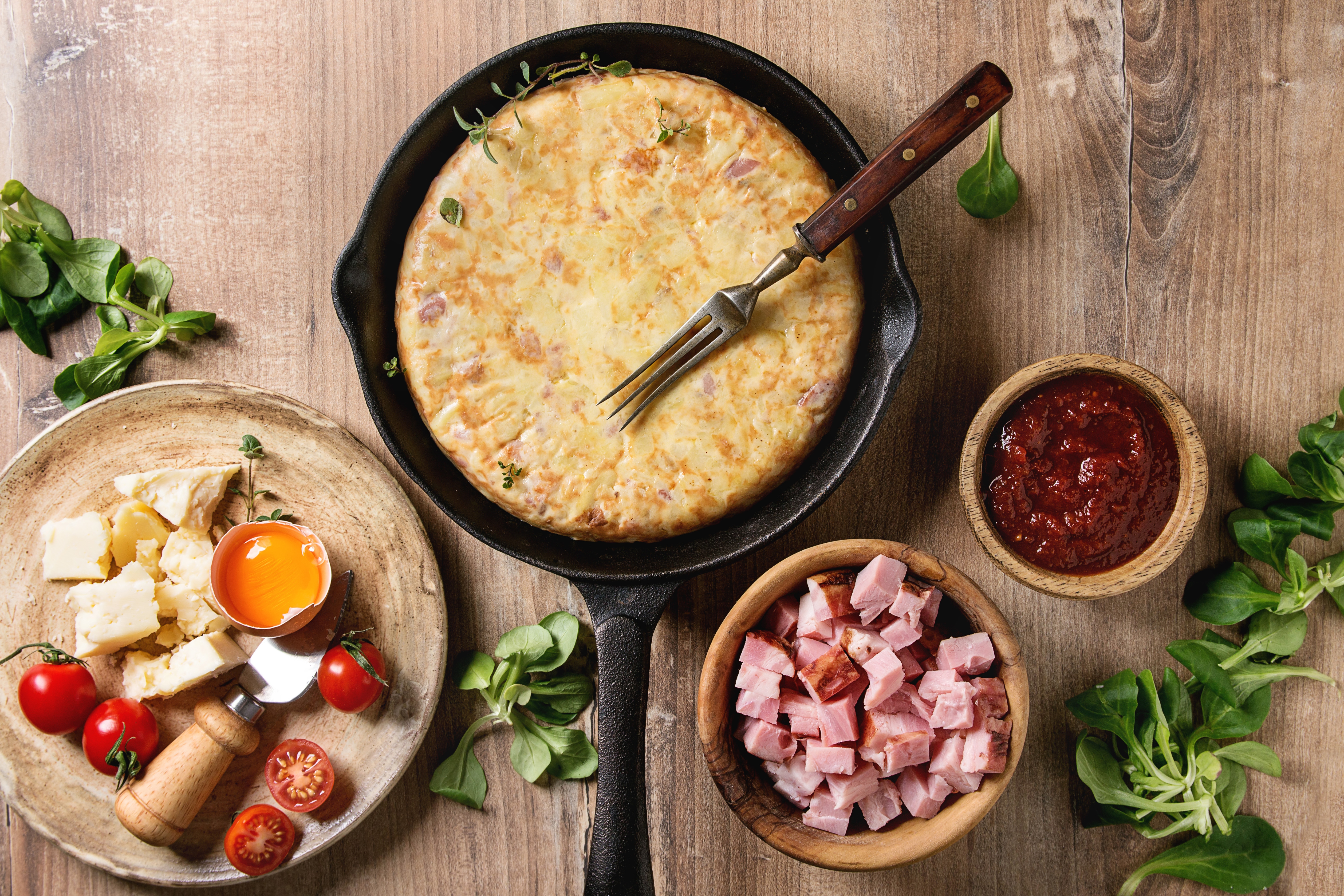 Spanish potato omelette tortilla with bacon served in cast-iron pan with sauce and ingredients above over wooden background Top view, space