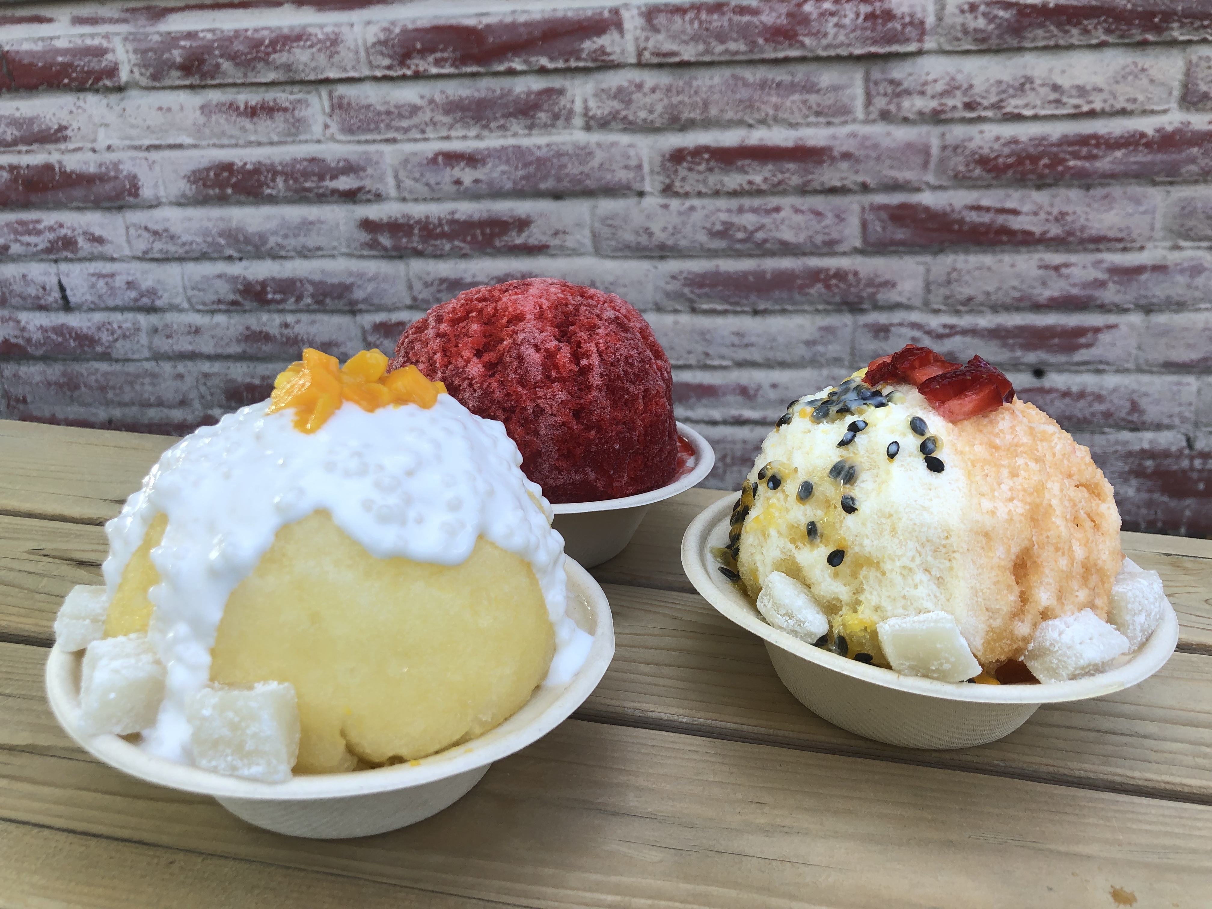 Three bowls of shave ice on a light wood table in front of a red brick wall. 