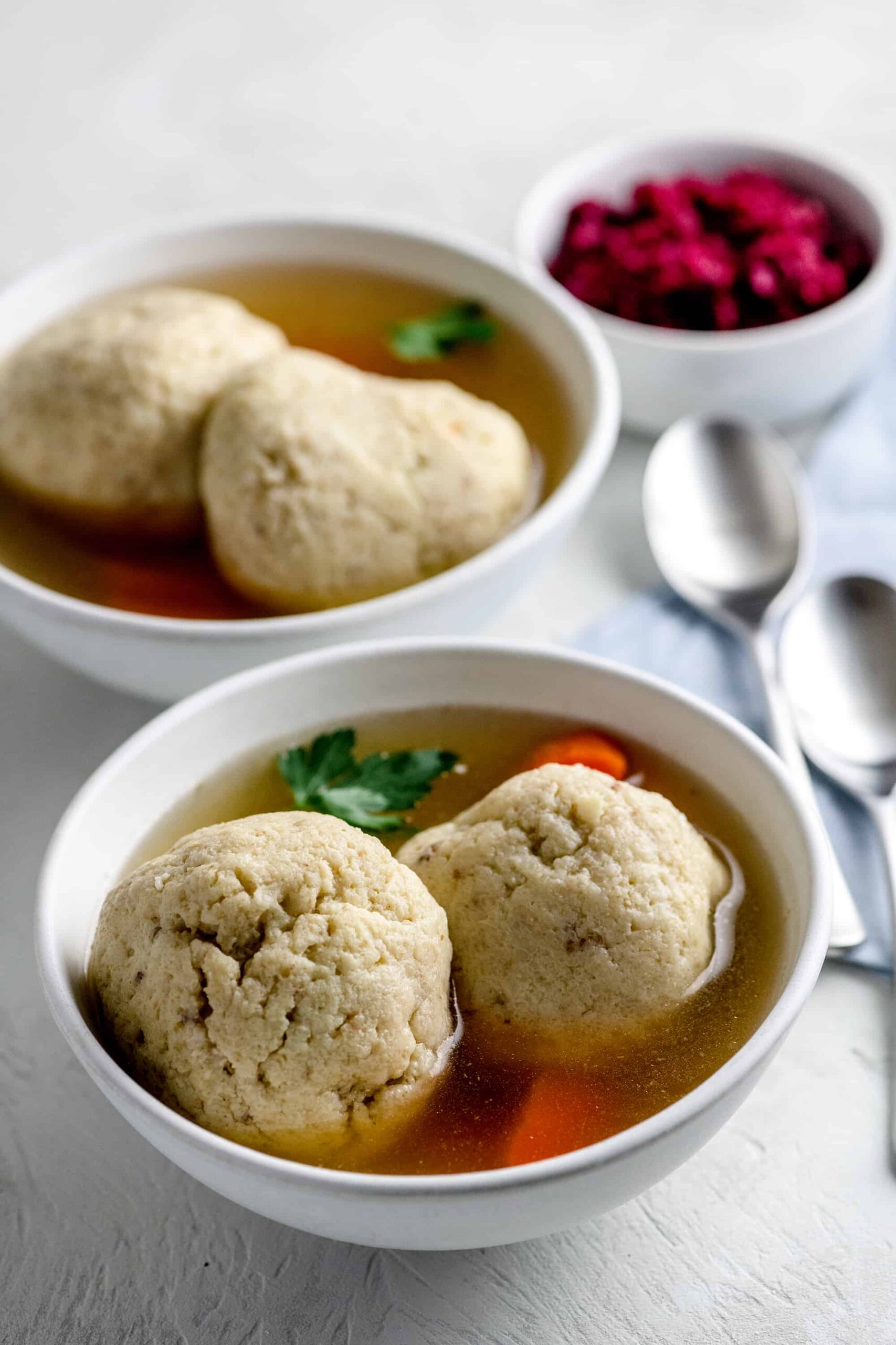 Two bowls of matzo ball soup are served on a white tablecloth with silver spoons.