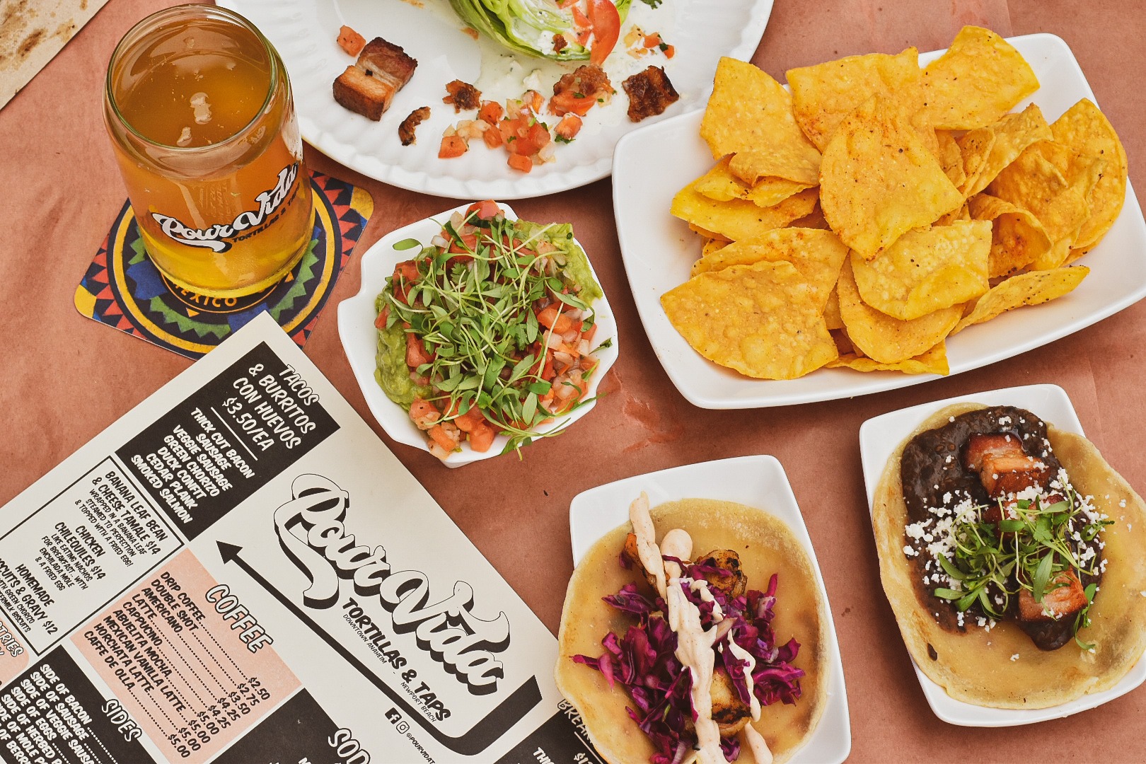 An overhead shot of tacos, chips, and a beer and menu.
