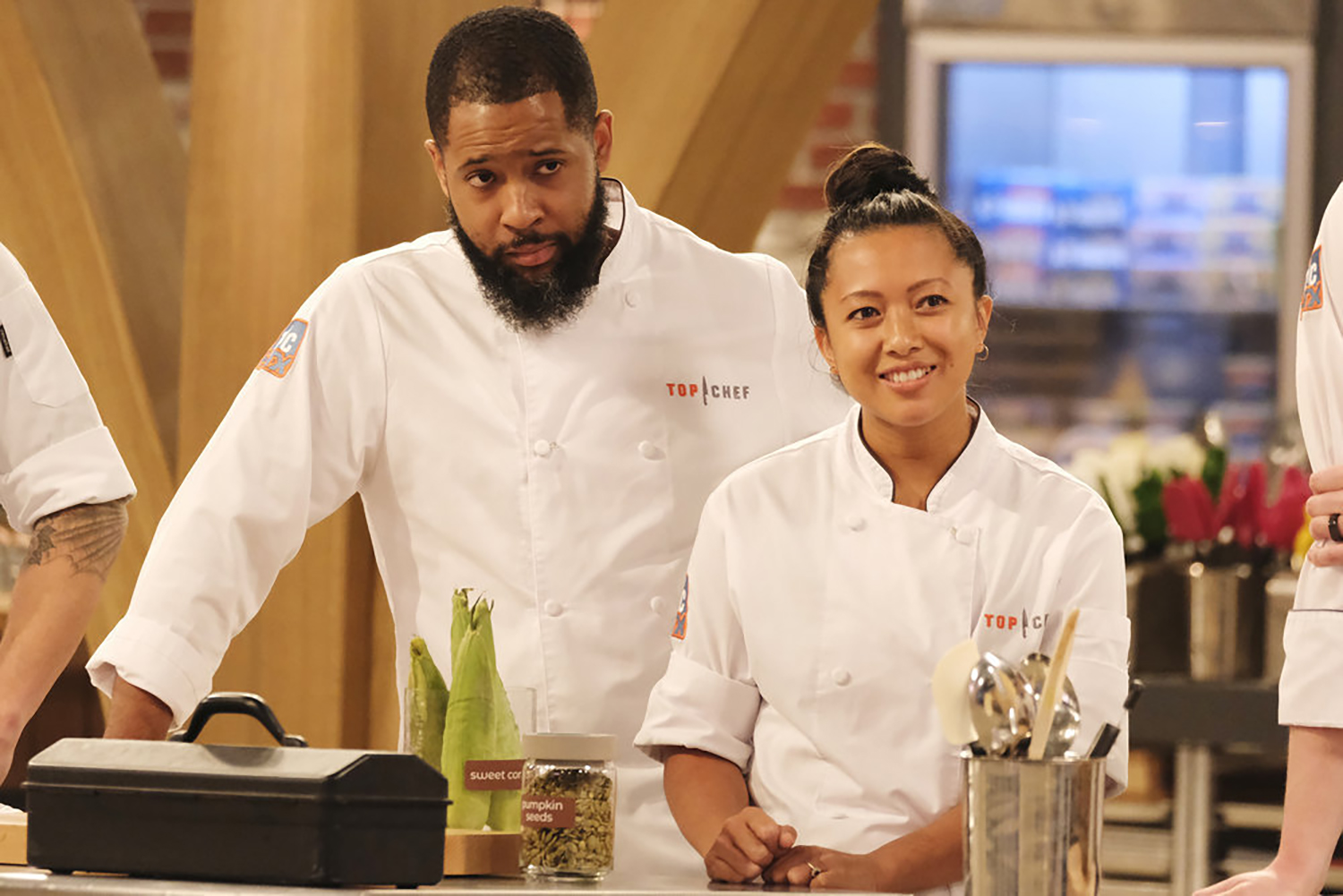 A male chef and a female chef in jackets stand behind a cooking counter listening to someone off camera.