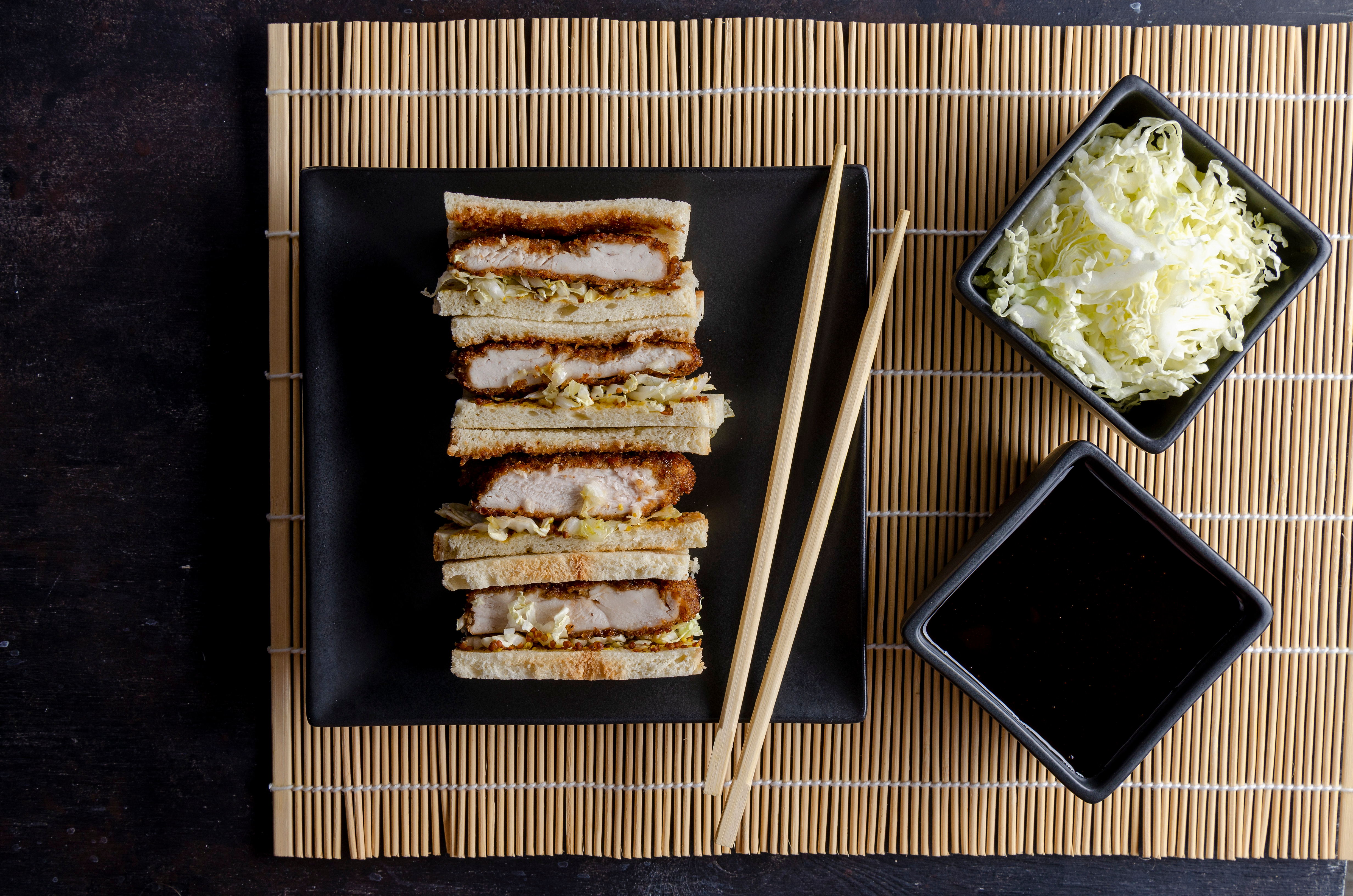 A stock image of four katsu sandwiches, layered with cabbage, with a side of katsu sauce and more shredded cabbage.