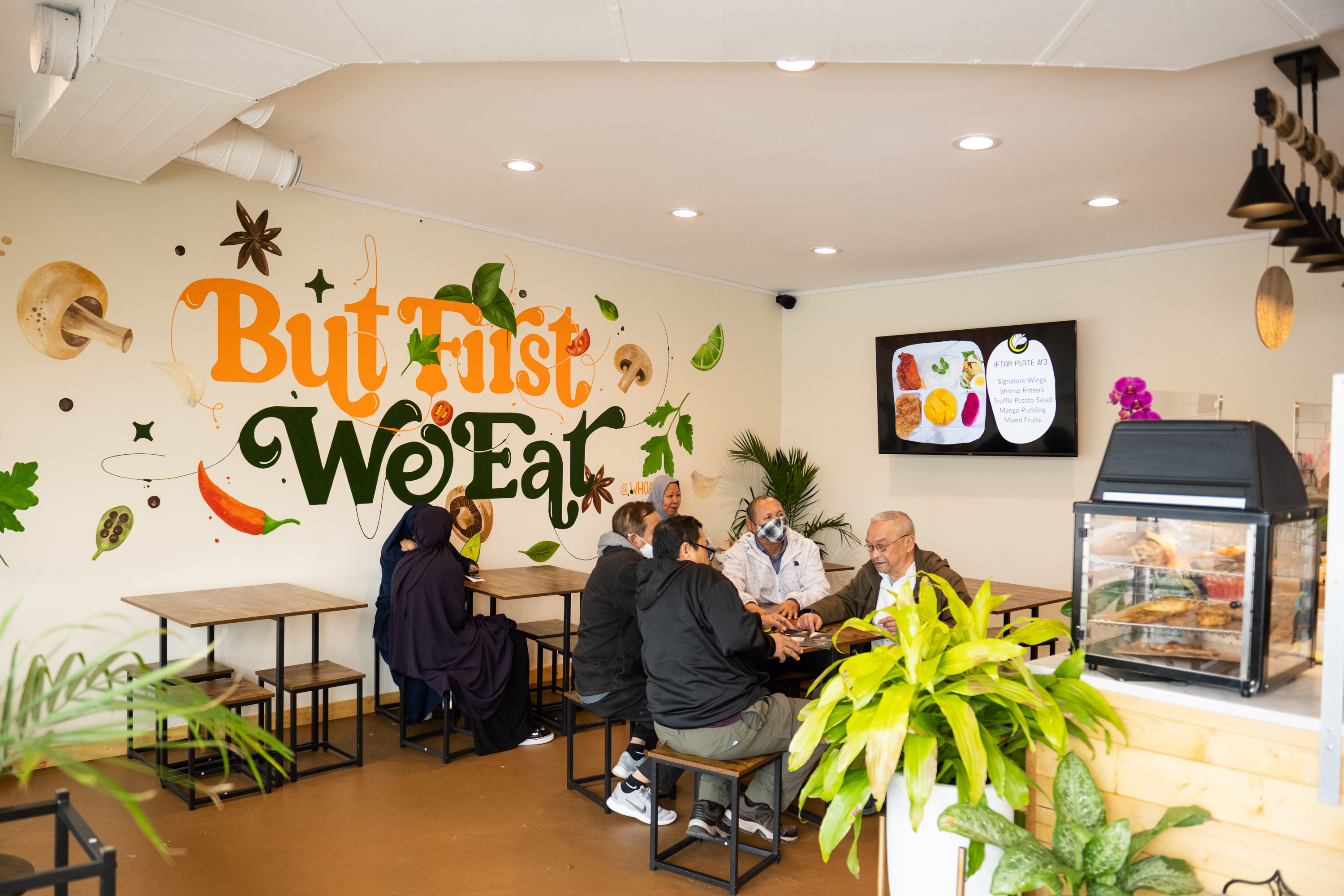 People dine at tables under a large mural that says, “first, we eat,” in a brightly lit restaurant with lots of plants and a wood floor. 