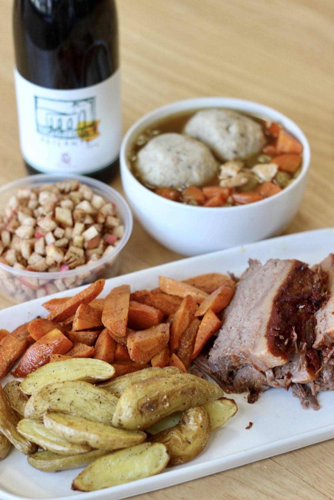 A white rectangular plate loaded with sliced brisket and roasted carrots and potatoes with a white ceramic bowl full of matzo ball soup behind it along with a takeout container full of chariest and a bottle of wine.