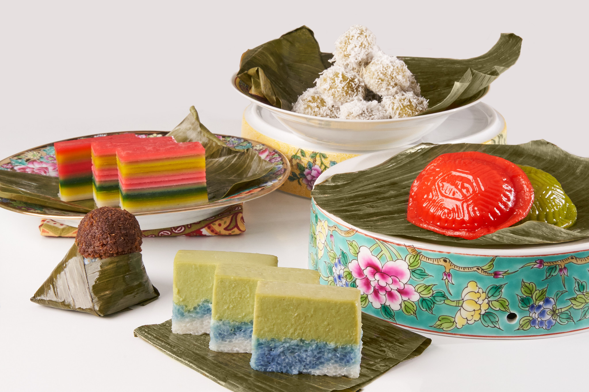 An array of colorful desserts laid out on green banana leaves and patterned platters.