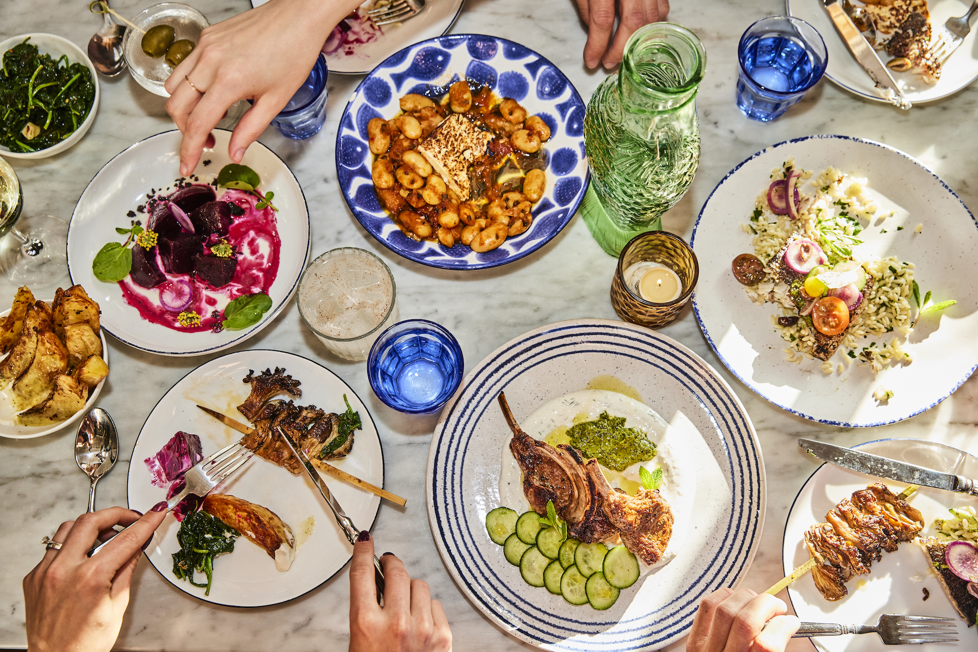 A marble table filled with plates of Greek food like grilled meats, as hands reach in.