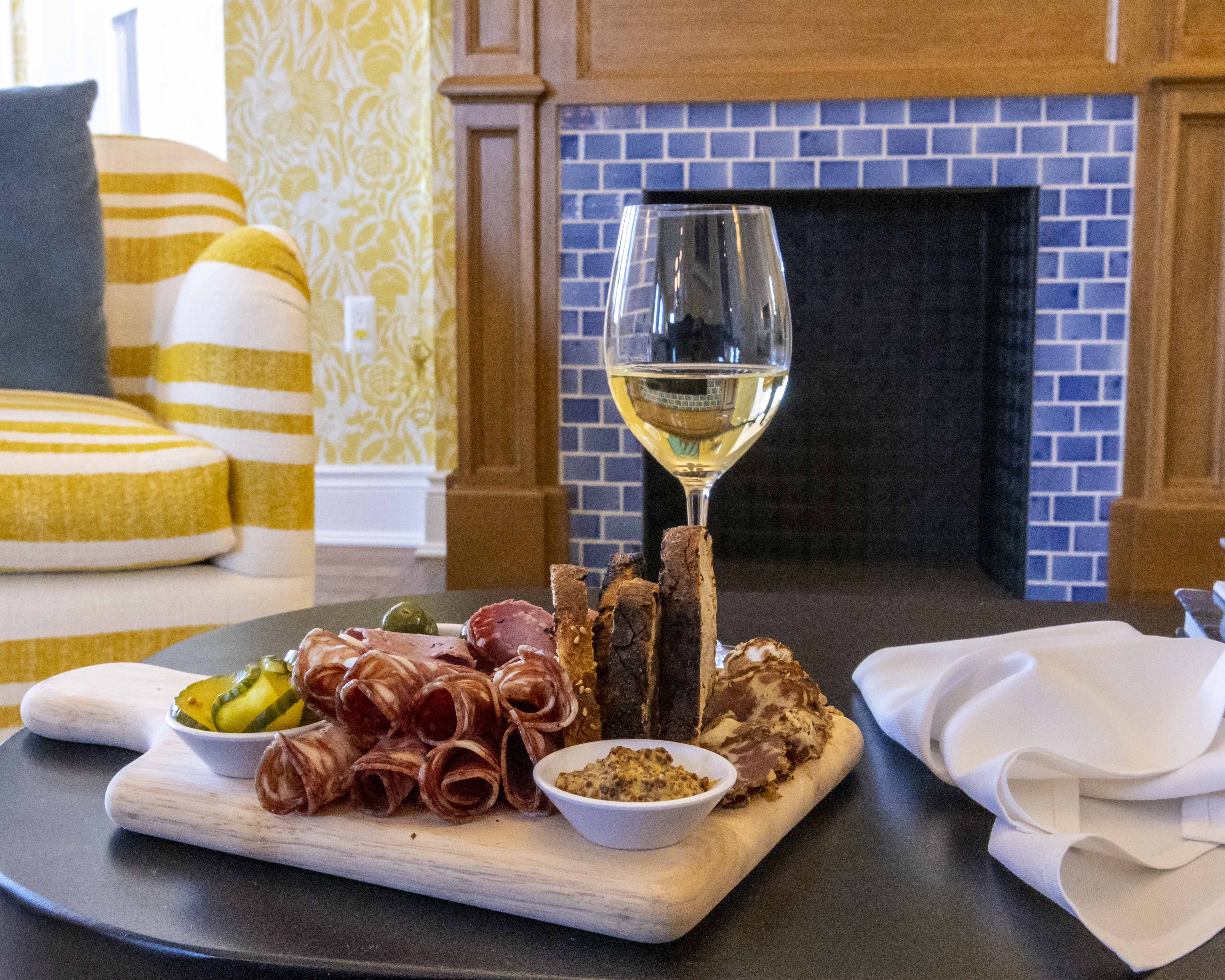 A charcuterie board with meat, veggies, bread, and dip on a wooden cutting board on a black round top table, with a blue tiled and wooden fireplace and yellow and white upholstered chain with a blue pillow