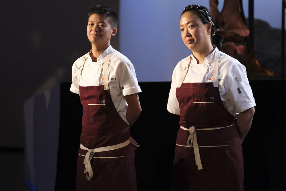 Two women in chef’s garb and red aprons standing in a dramatically lit room. 