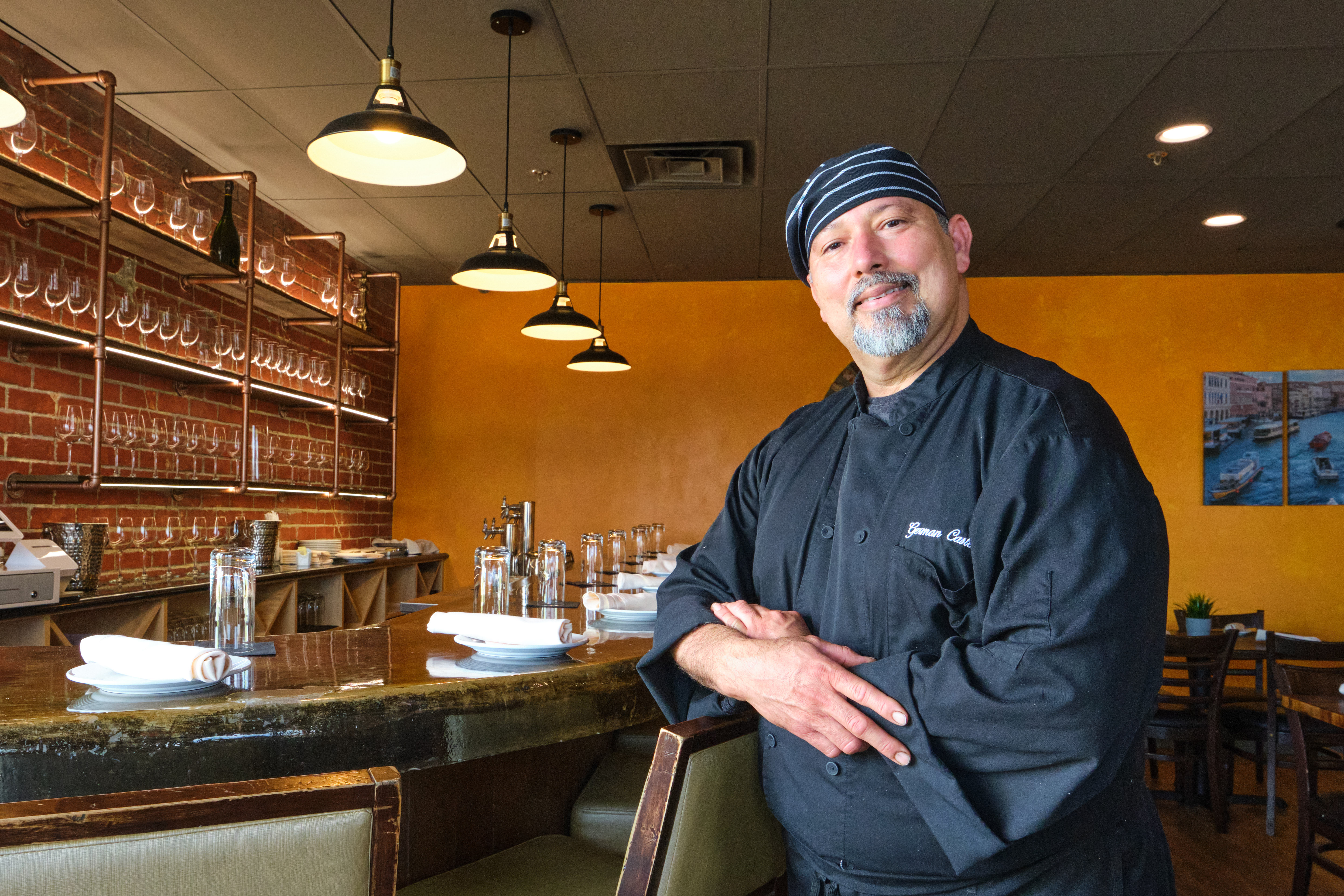 A chef in a black coat poses in front of a bar.