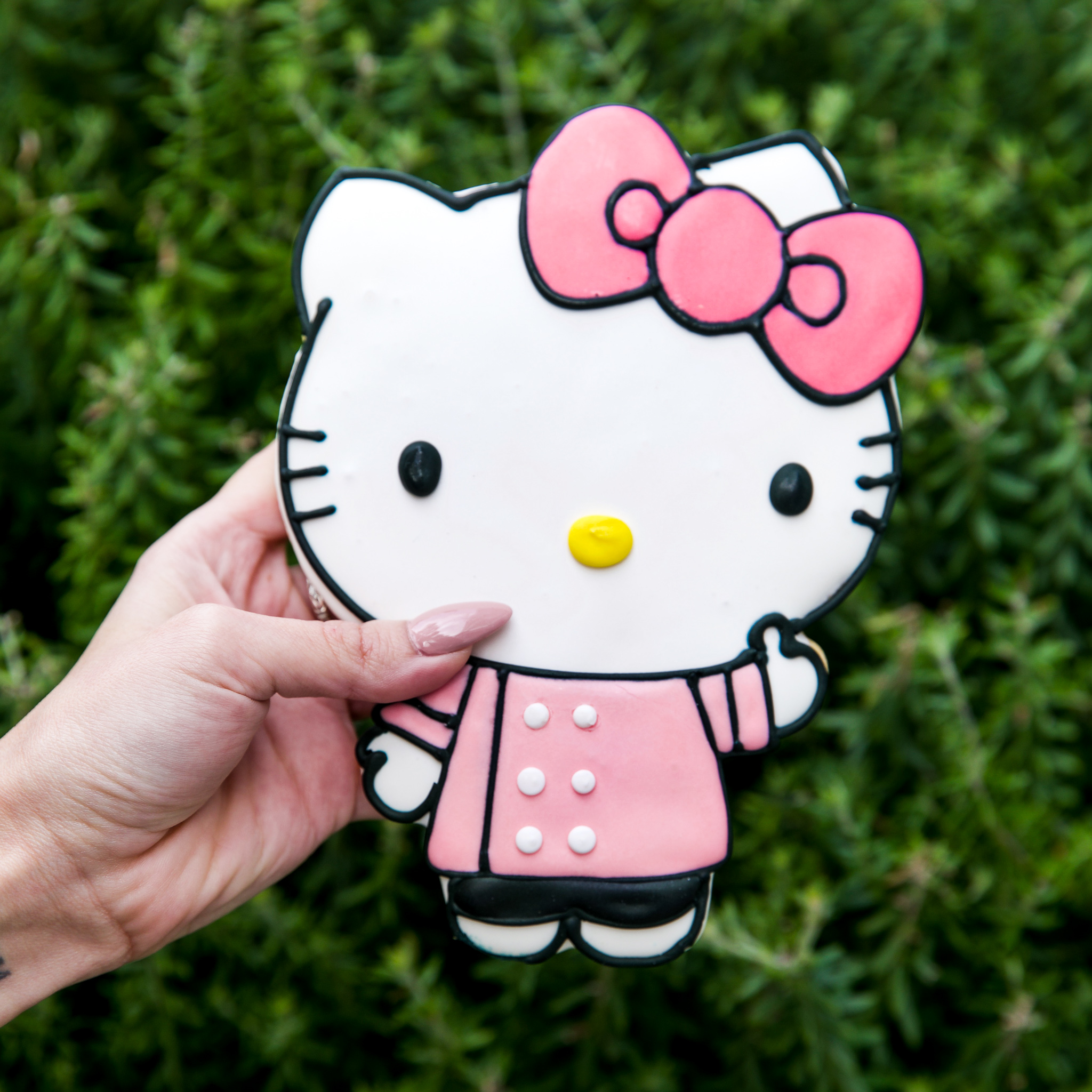 A white woman’s hand with long pink nails holds an oversized Hello Kitty cookie. Hello Kitty wears a pink smock top and a matching pink bow, with a yellow nose, white face and black whiskers.