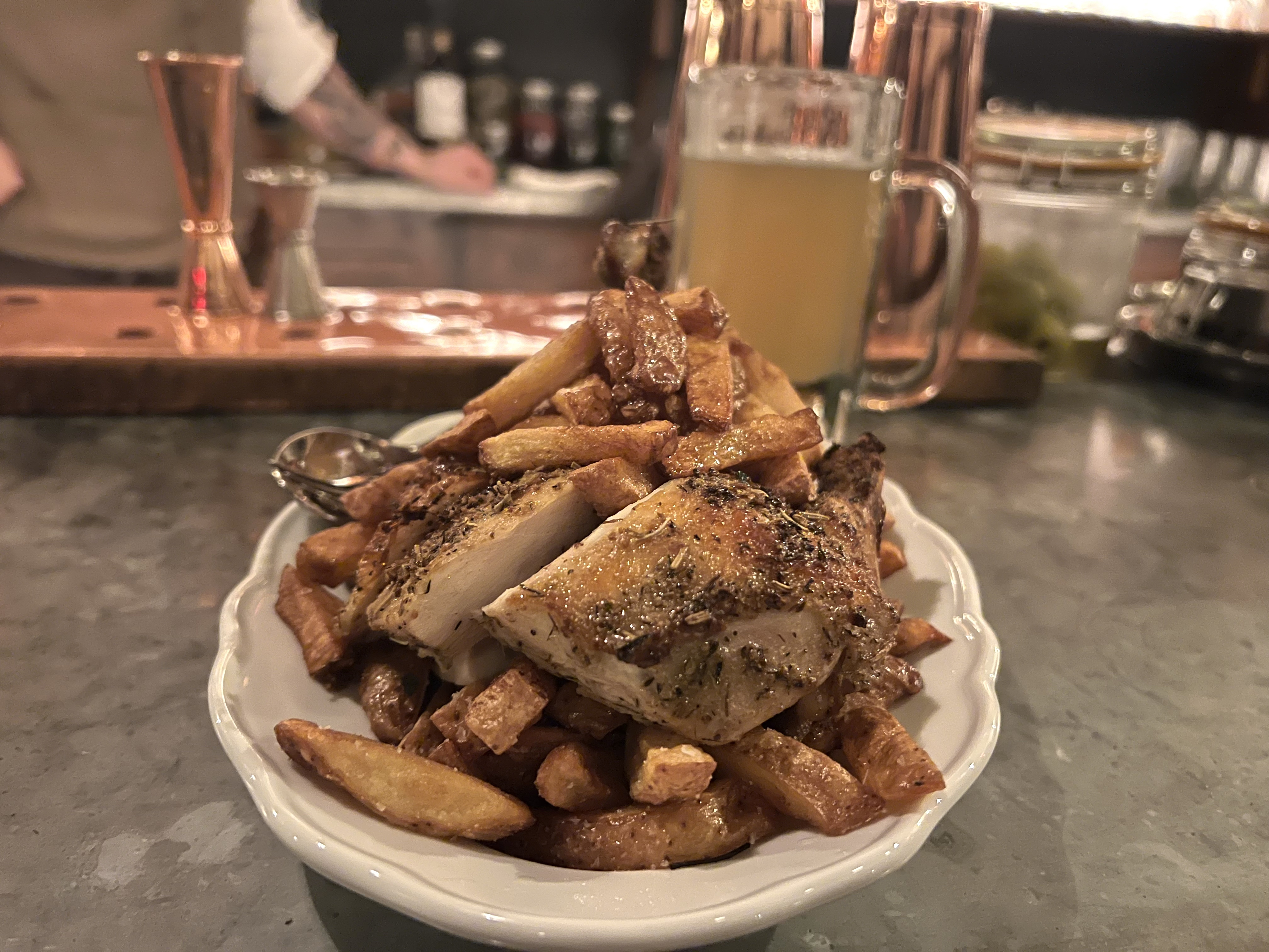 Roast chicken, herbed and bronzed, sits over a pile of fries on a white plate. A glass mug of beer is visible behind the dish.