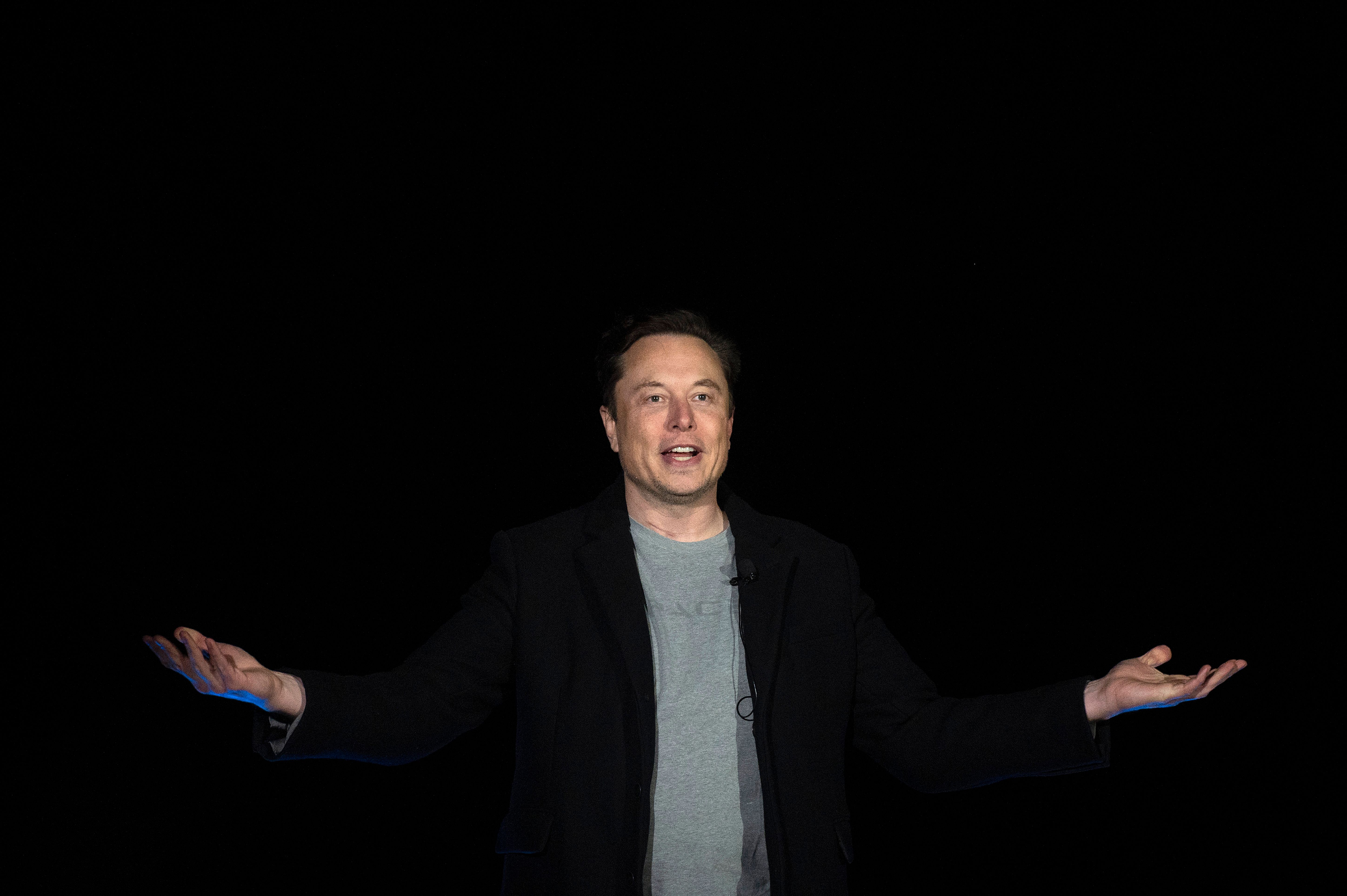 Elon Musk standing against a dark background with his hands out in a “who me?” gesture.