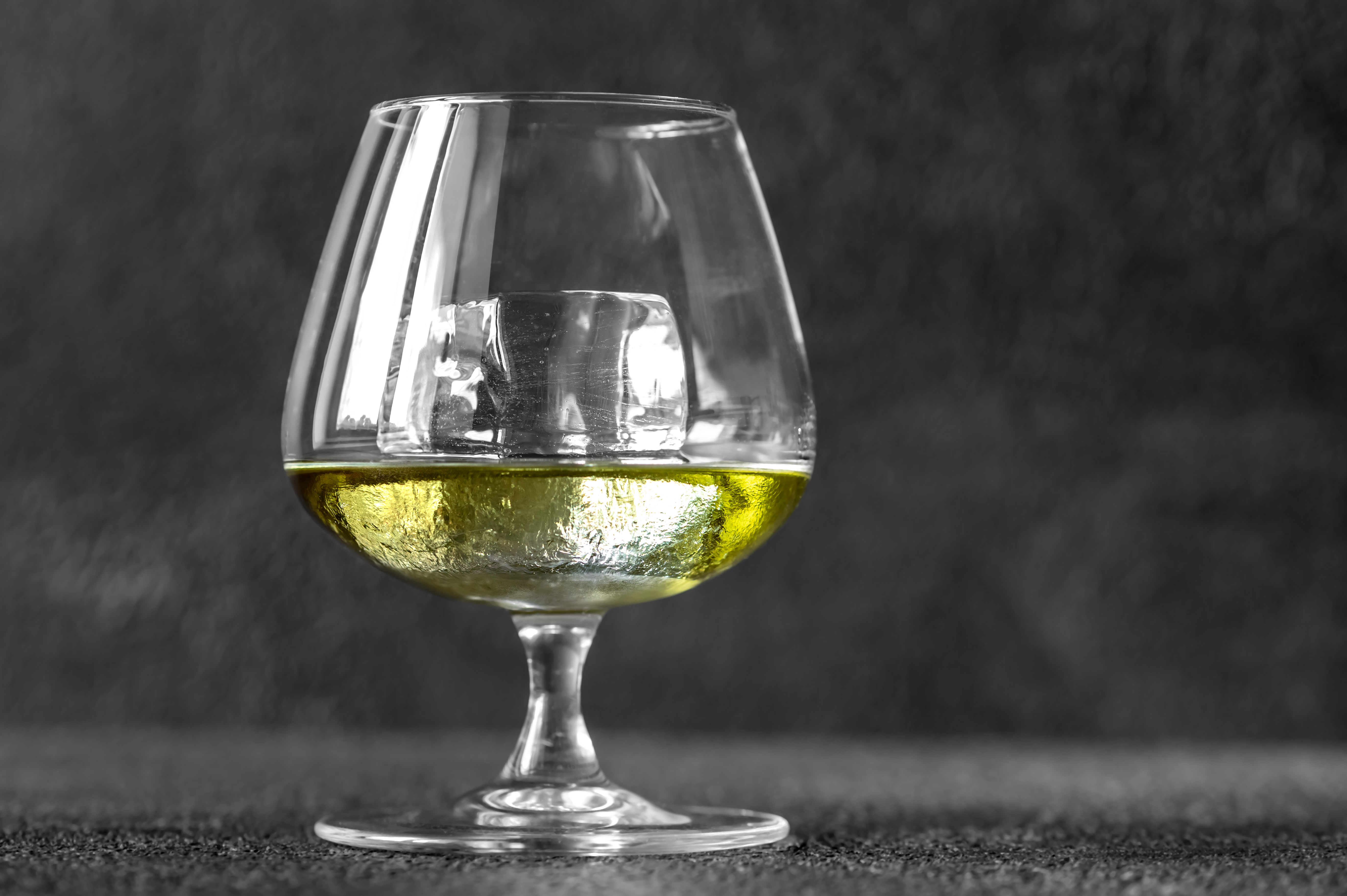 A glass of chartreuse with a single ice cube against a dark gray background. 