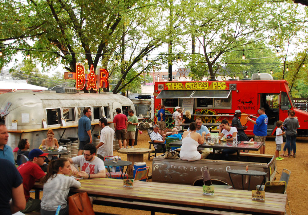Customers sit at wooden picnic tables on a dirt patio at Truck Yard in Lower Greenville. To the left is a silver Airstream with a red sign reading “bar” and to the right is a red food truck called The Crazy Pig