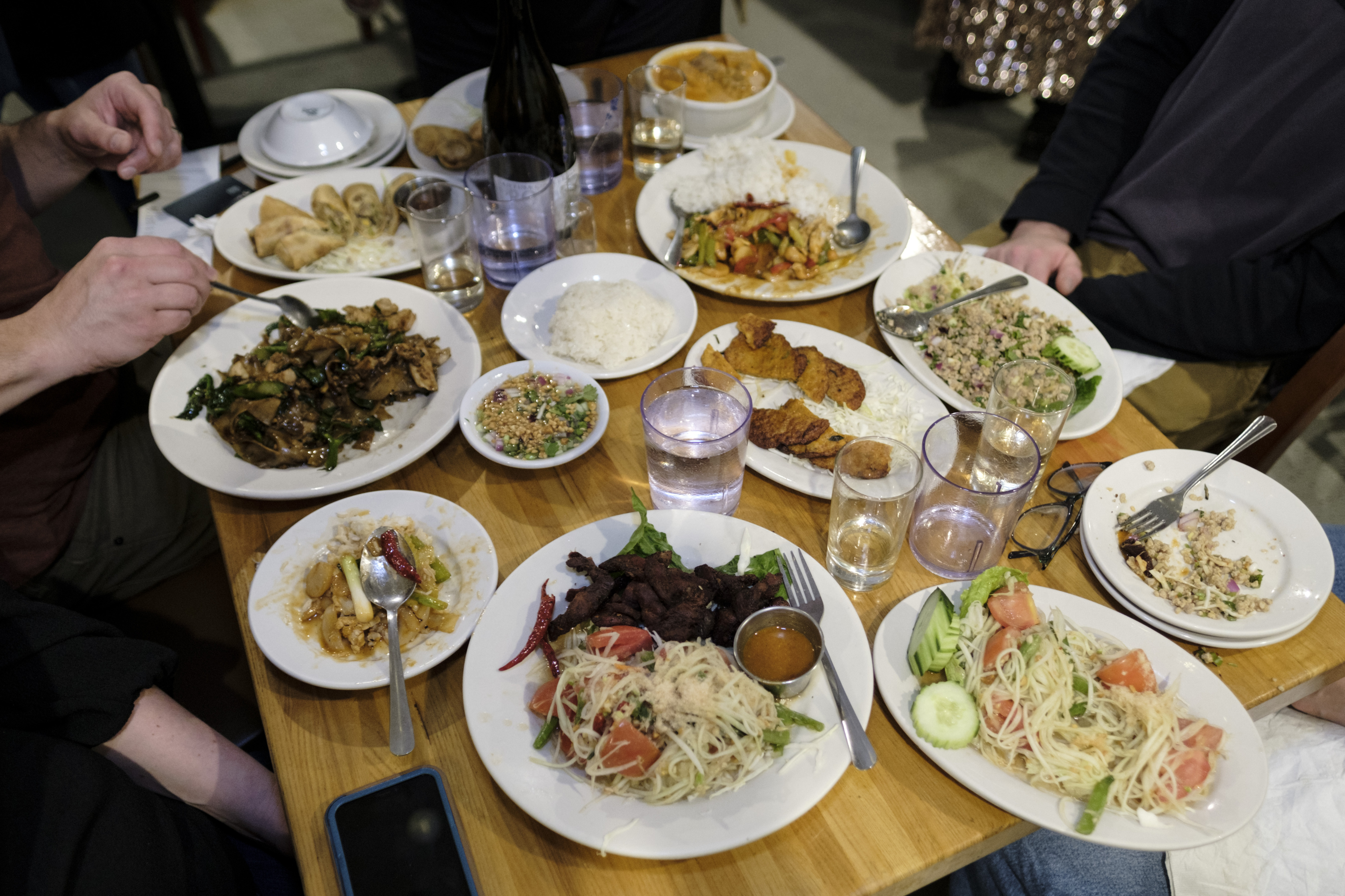 A wood table is set with various Thai dishes on white plates: green papaya salad, fried pork, fish cakes, and other rice and noodle dishes.