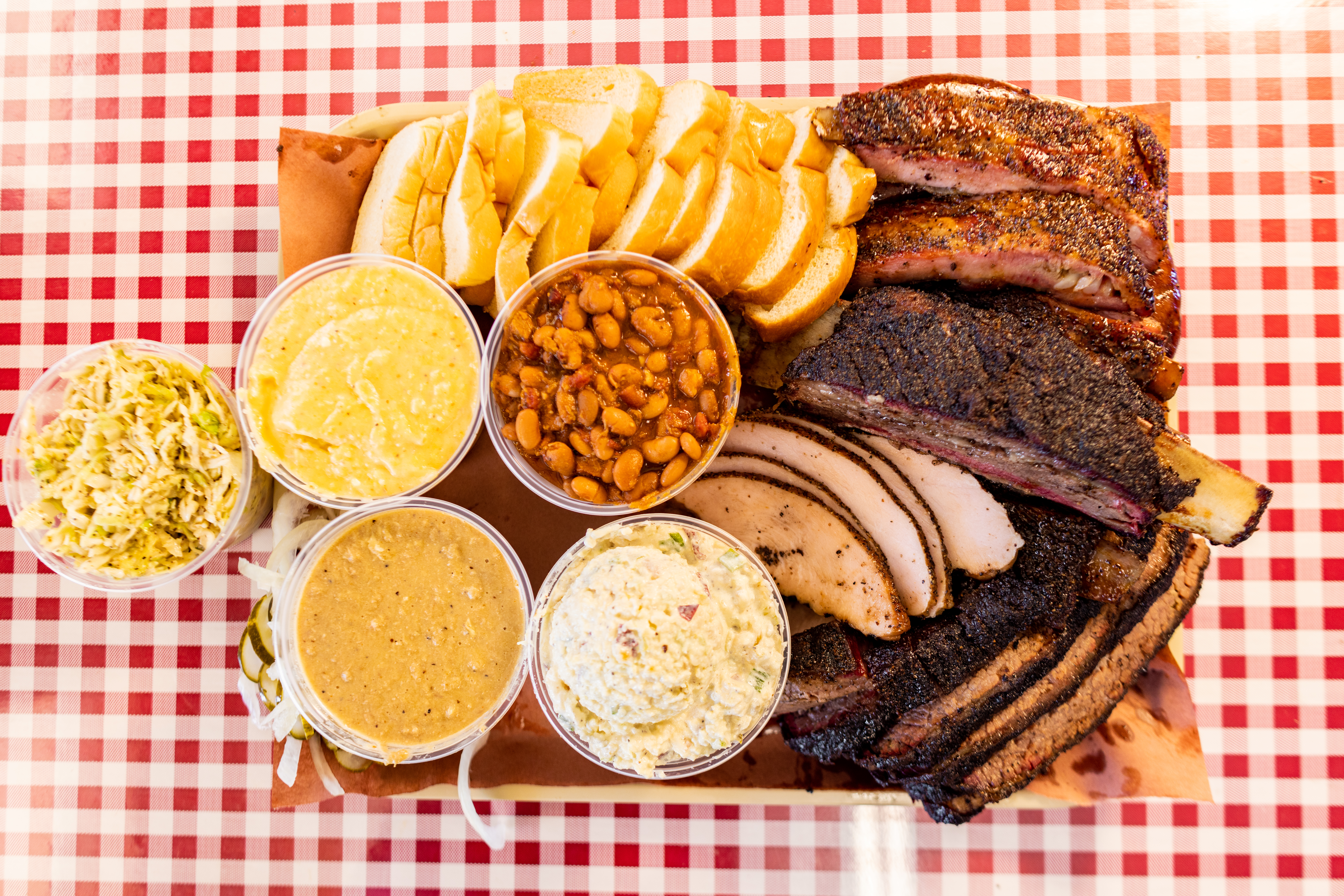 A platter of ribs, brisket, turkey, sliced bread, beans, potato salad, and other sides on a red and white checked tablecloth. 