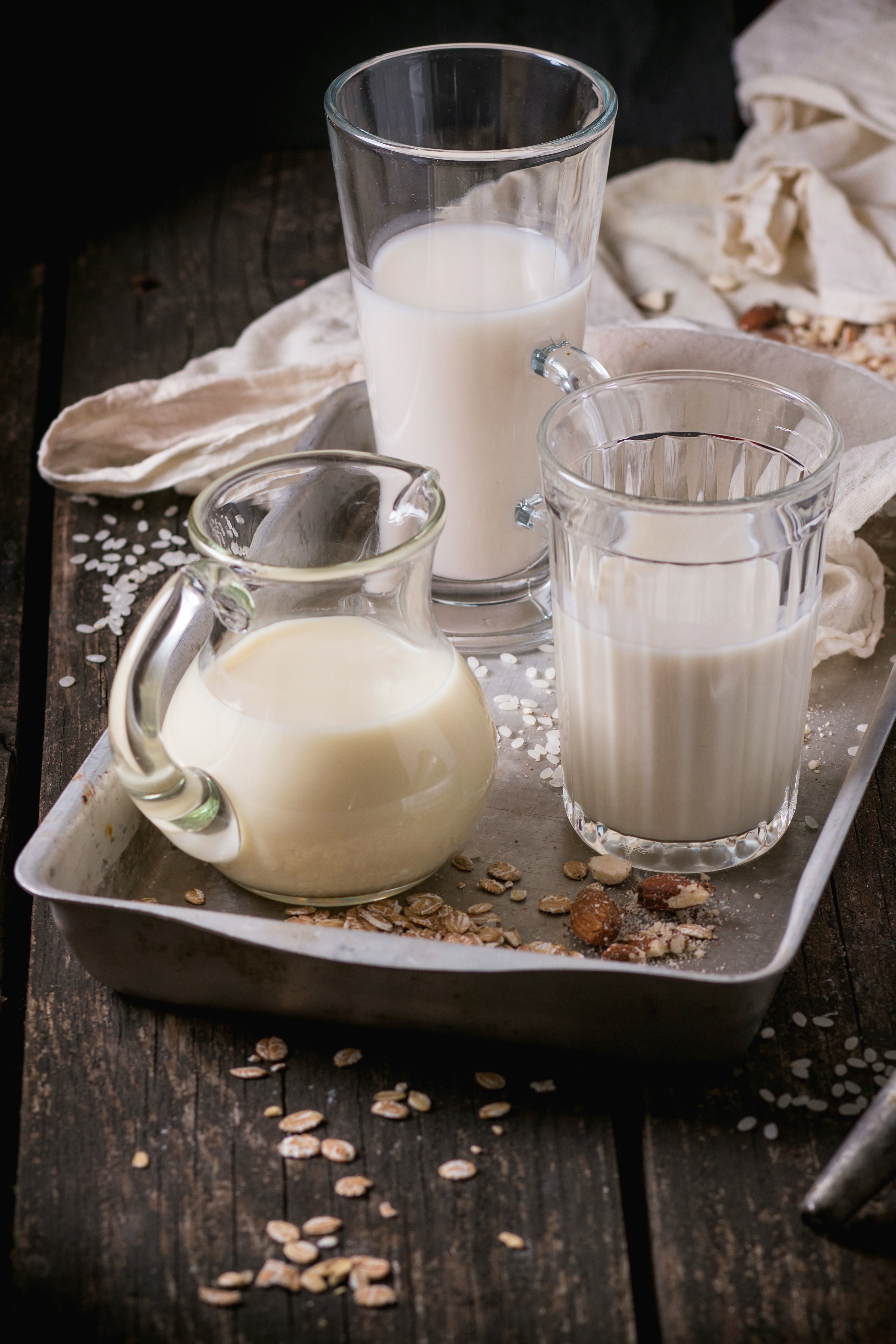 Set of non-dairy milk (rice milk, almond milk and oat milk) in glass cups and jug on old aluminum tray with rice grains, oat flakes and almond over old wooden table. Dark rustic style.