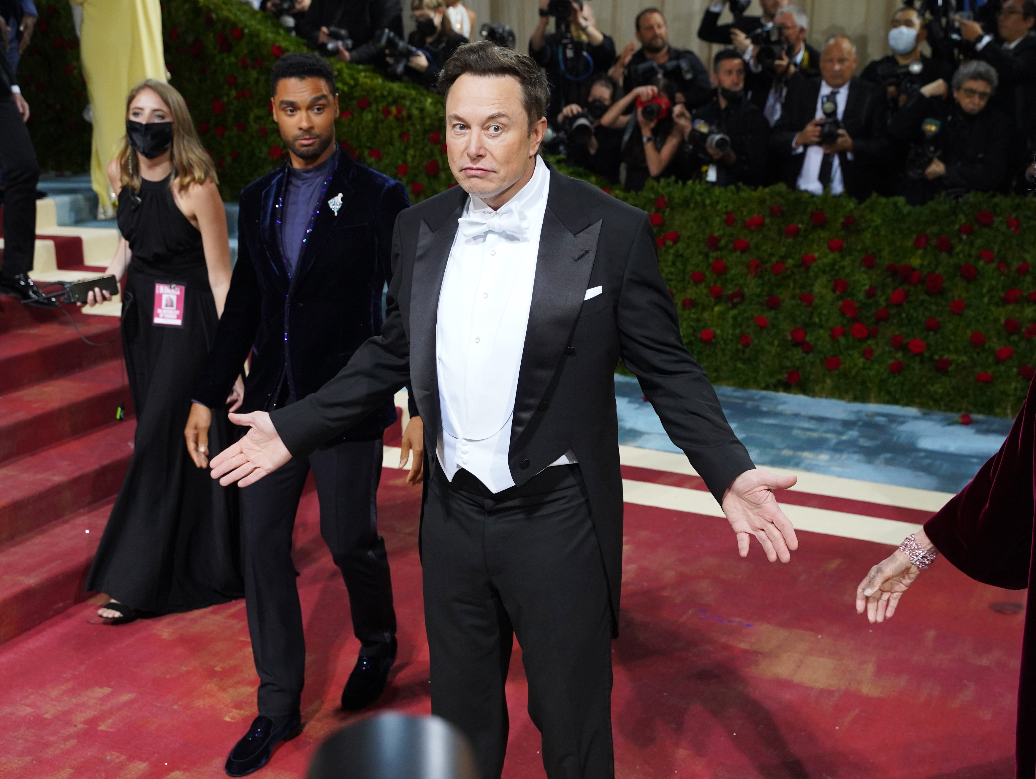 Elon Musk, wearing a tuxedo and making a “who me?” gesture, at a red carpet event.