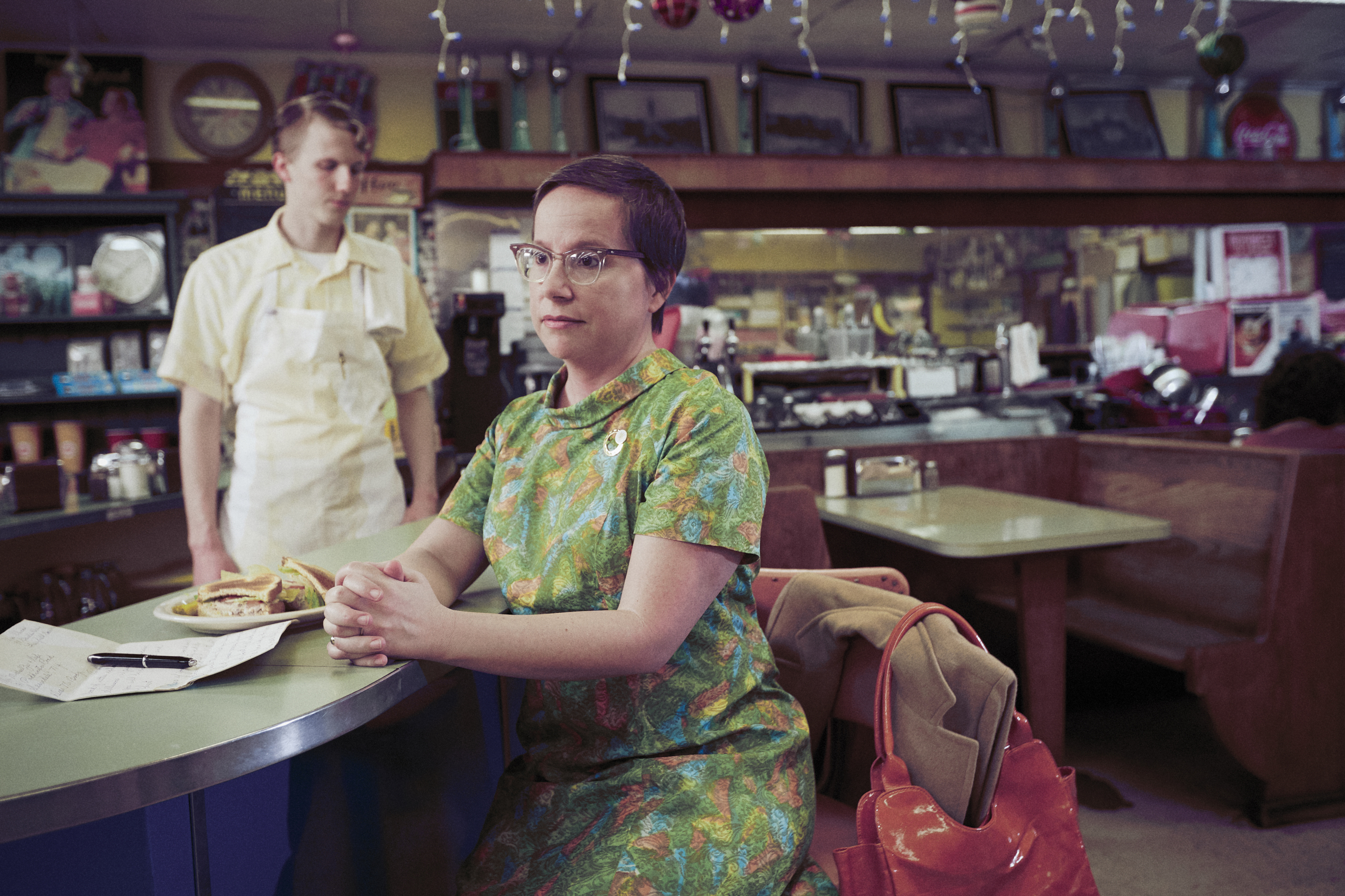 A woman in a green patterned dress and glasses sitting at a restaurant counter with a restaurant worker behind it.