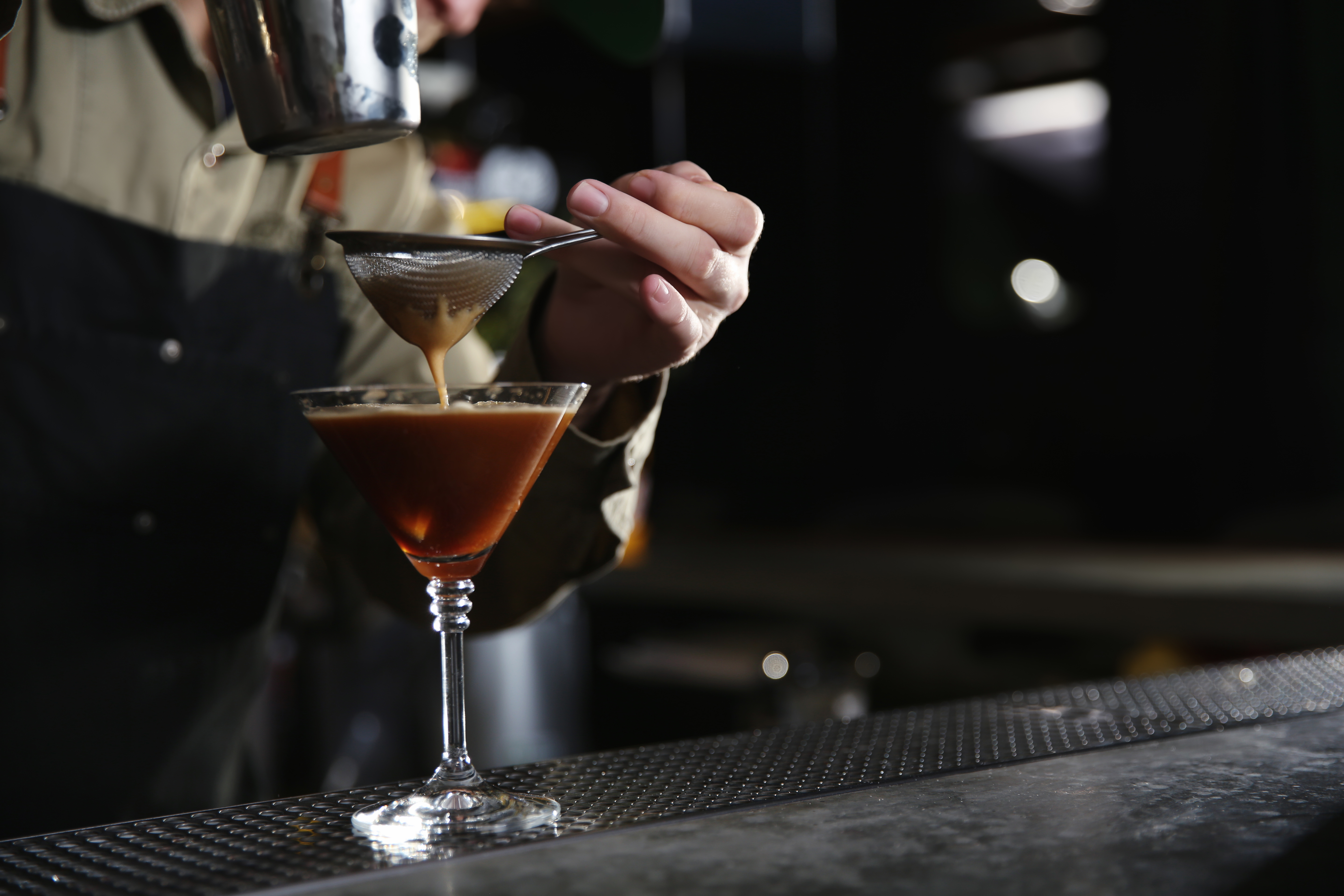 A bartender makes an espresso martini using a sieve at a bar.