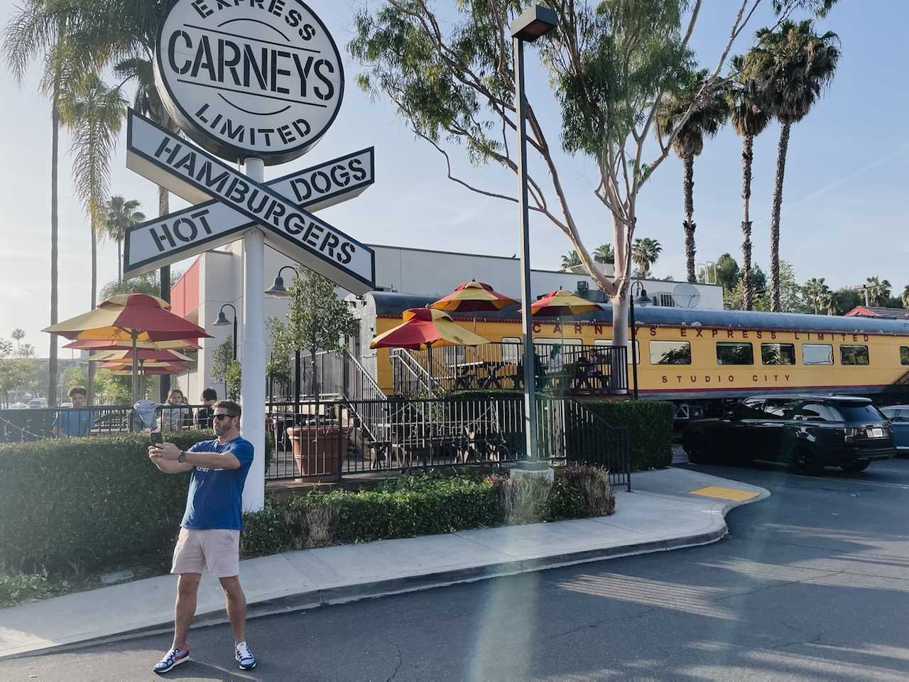 A man stands in front of Carney’s Restaurant while taking a selfie.