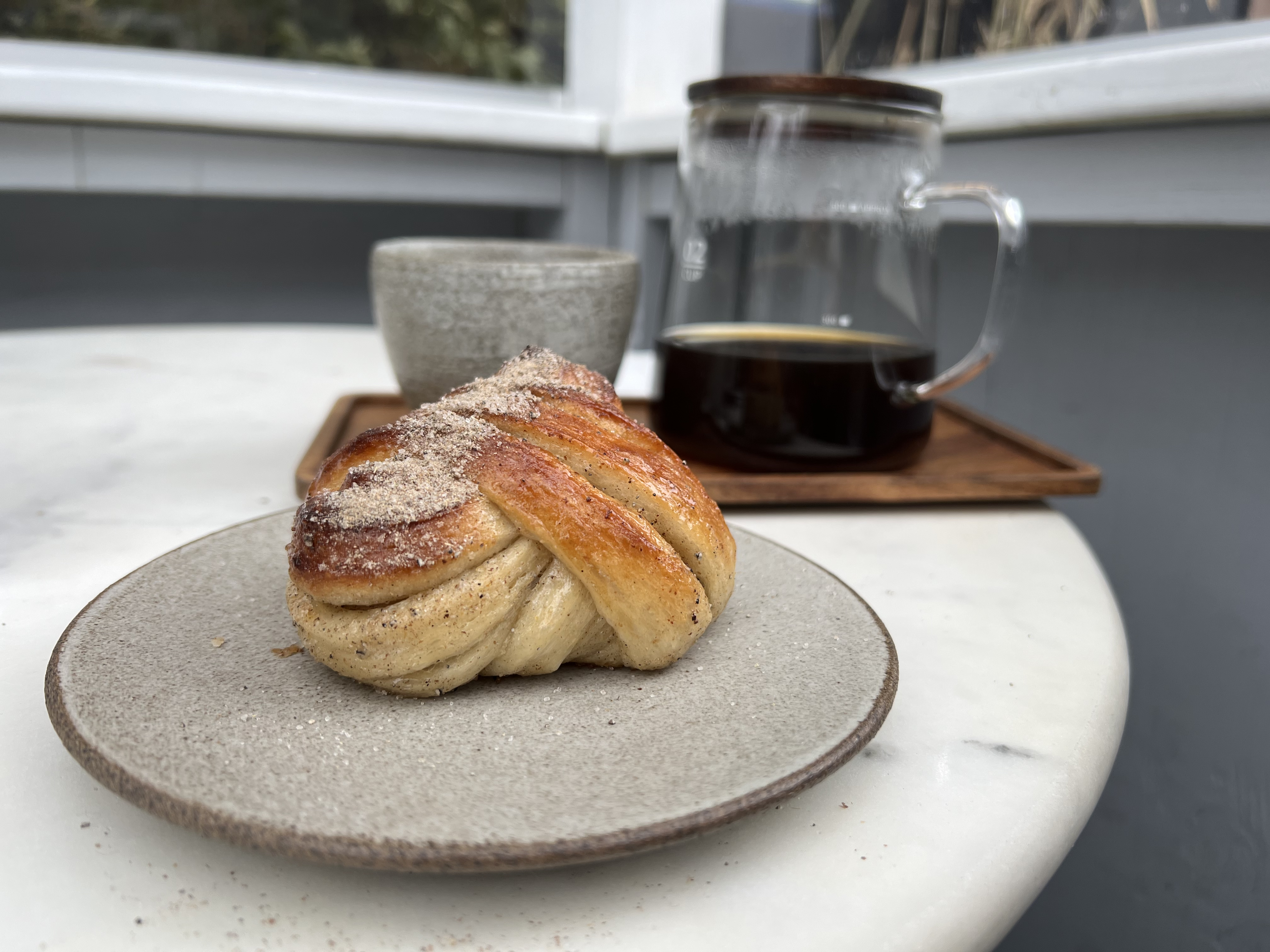 A cardamom bun sits on a grey plate on a white table, just in front of a clear coffee flask and an earthenware cup